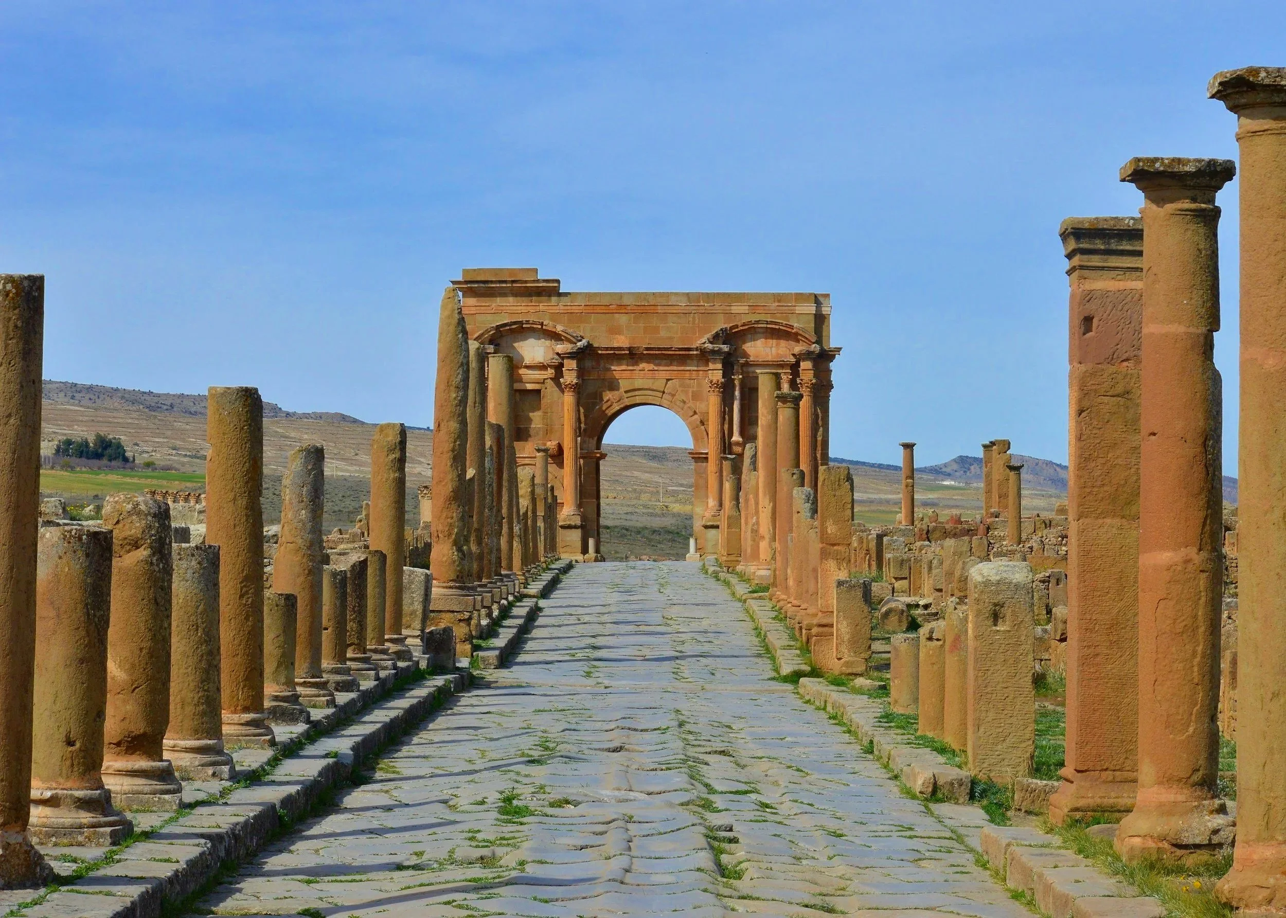 Ruinas de un antiguo camino con columnas y arcos en un paisaje abierto con colinas en el fondo.