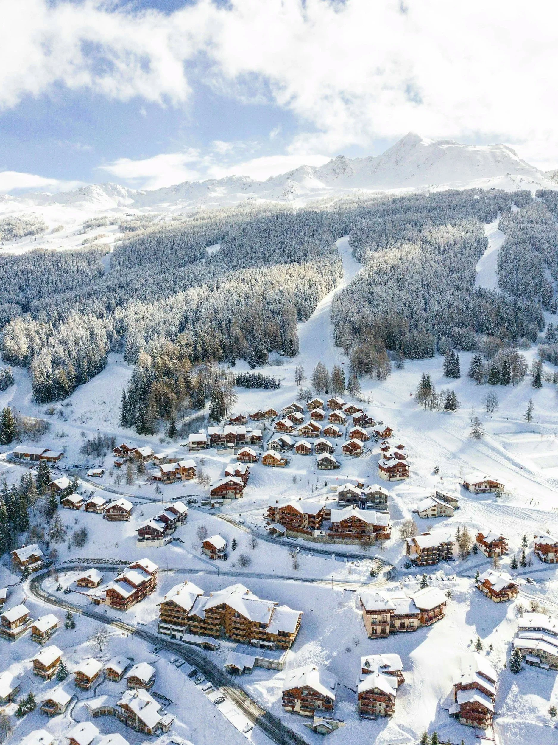 Vista aérea de un pueblo en la nieve rodeado de bosques y montañas con picos cubiertos de nieve y nubes en el cielo.