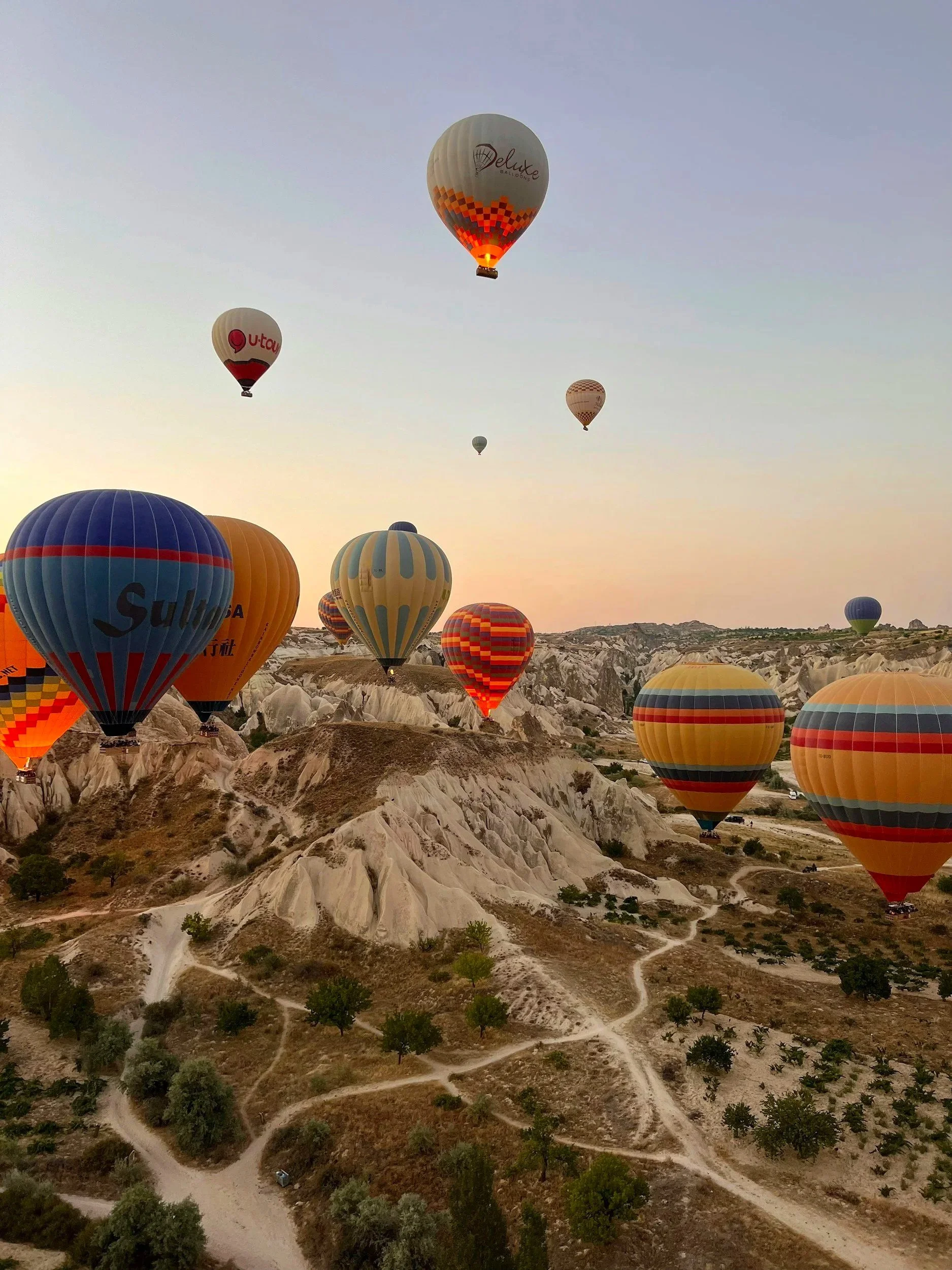 Balones de aire caliente flotando sobre un paisaje de formaciones rocosas y caminos en un amanecer o atardecer.