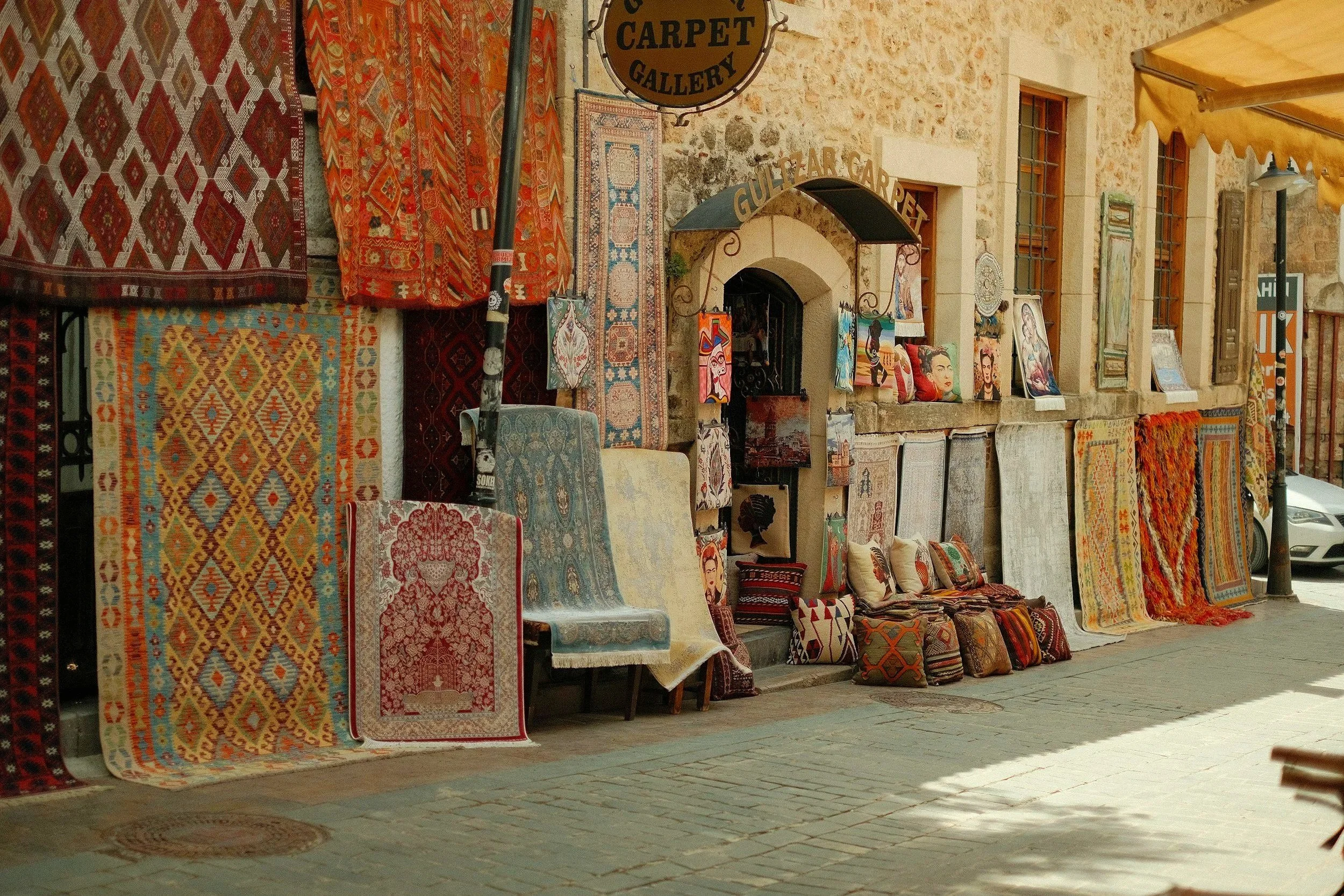 Carpeta, cojines y pinturas en una tienda de alfombras en una calle con pared de piedra y ventanas con barrotes.