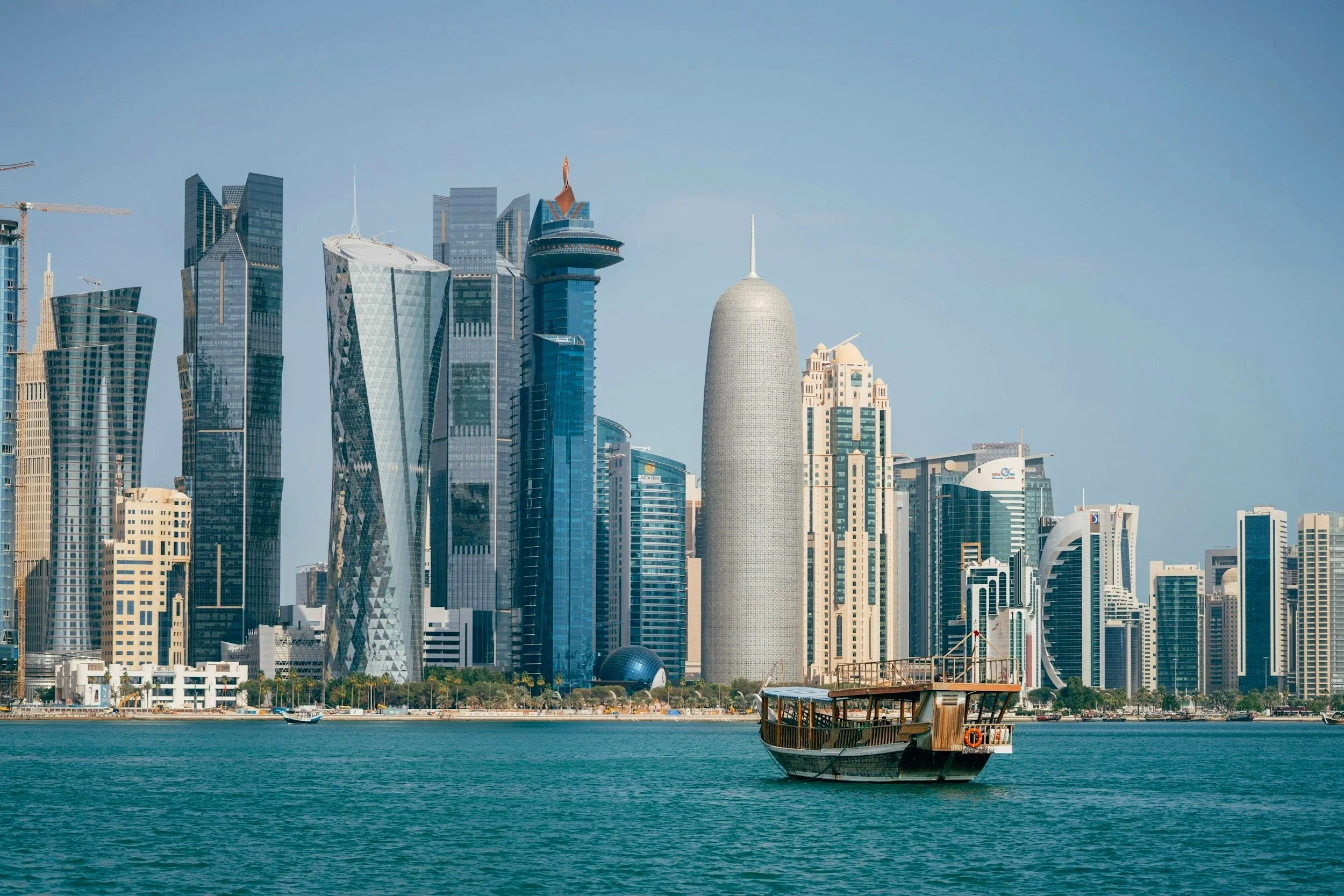 Vista del skyline de Doha, Catar, con rascacielos modernos y un barco tradicional en primer plano sobre el agua.