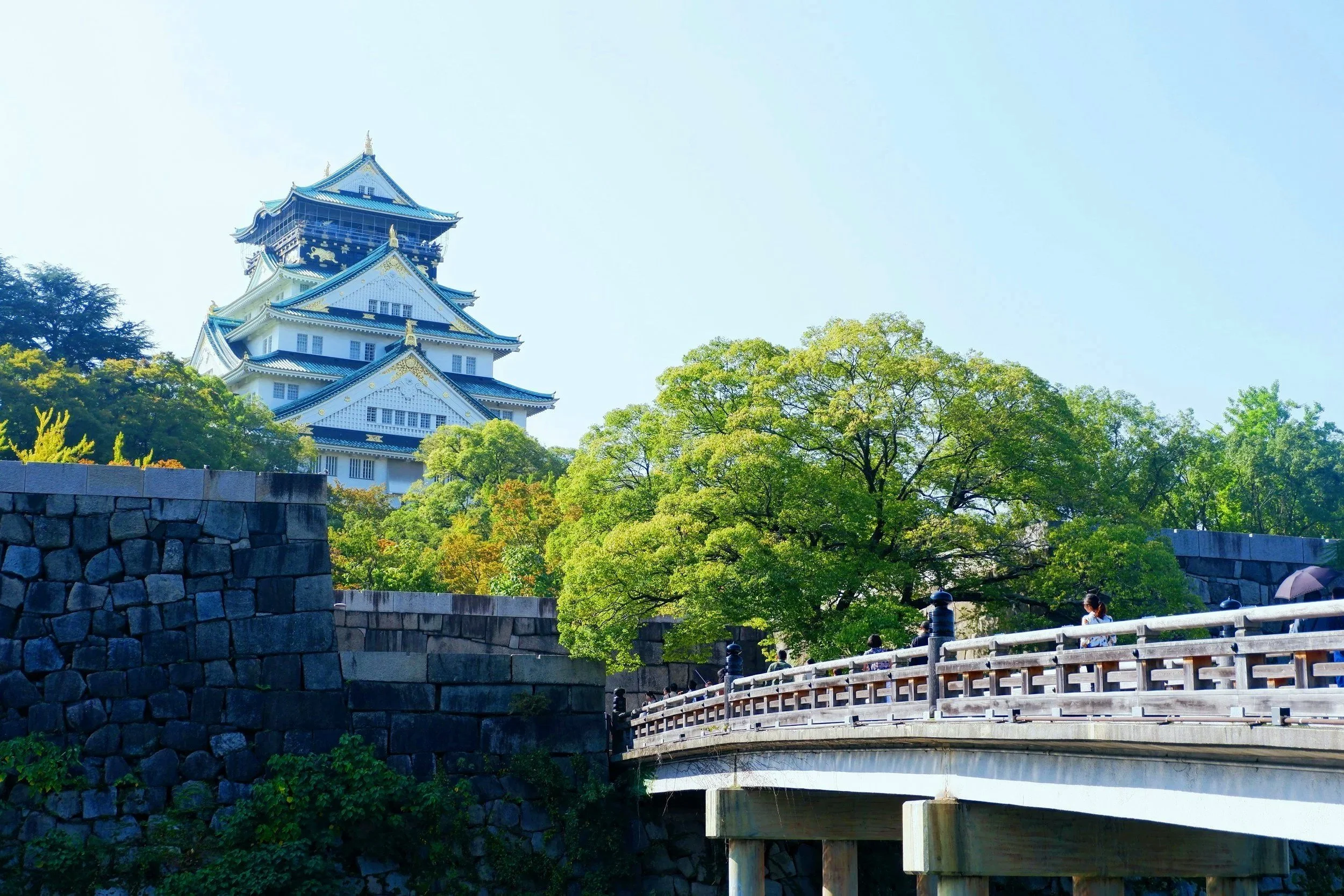 Castillo japonés sobre colina rodeado de árboles verdes, con puente de madera y personas paseando.