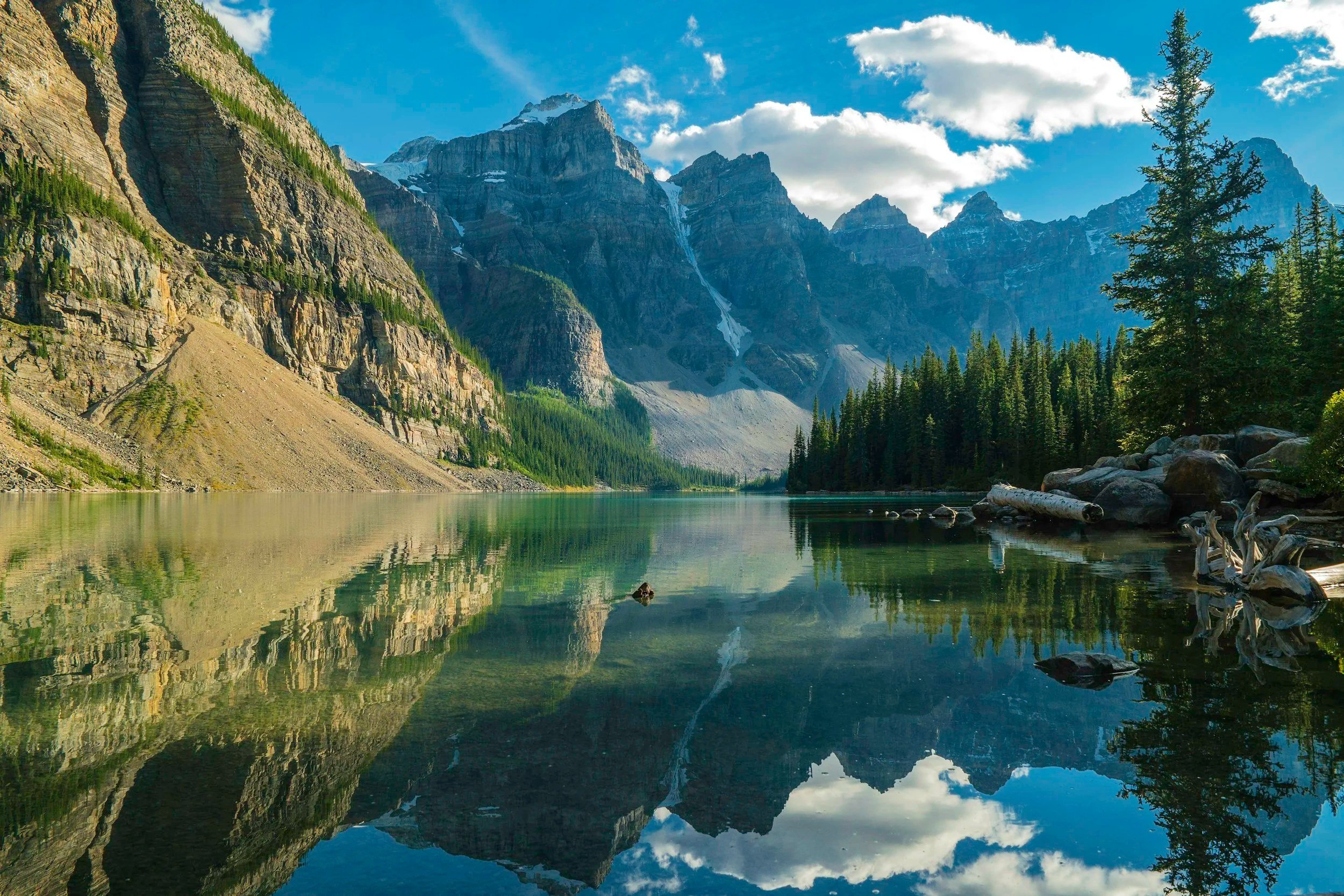 Paisaje de un lago rodeado de montañas con picos nevados y un bosque de árboles verdes, bajo un cielo azul con nubes.