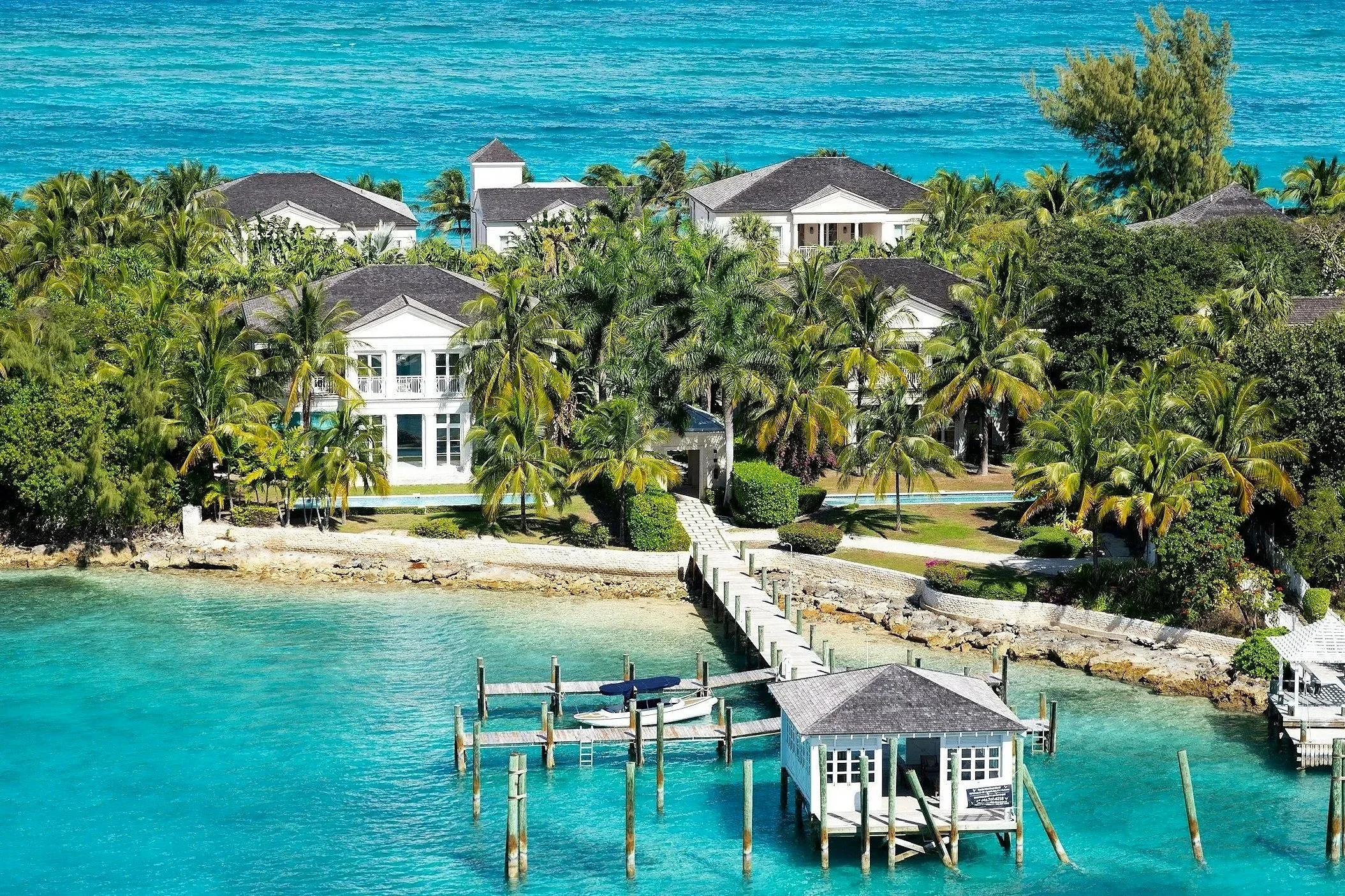 Casas blancas con techos grises, rodeadas de palmeras, frente a un mar azul y un muelle con cabañas, en un entorno tropical.