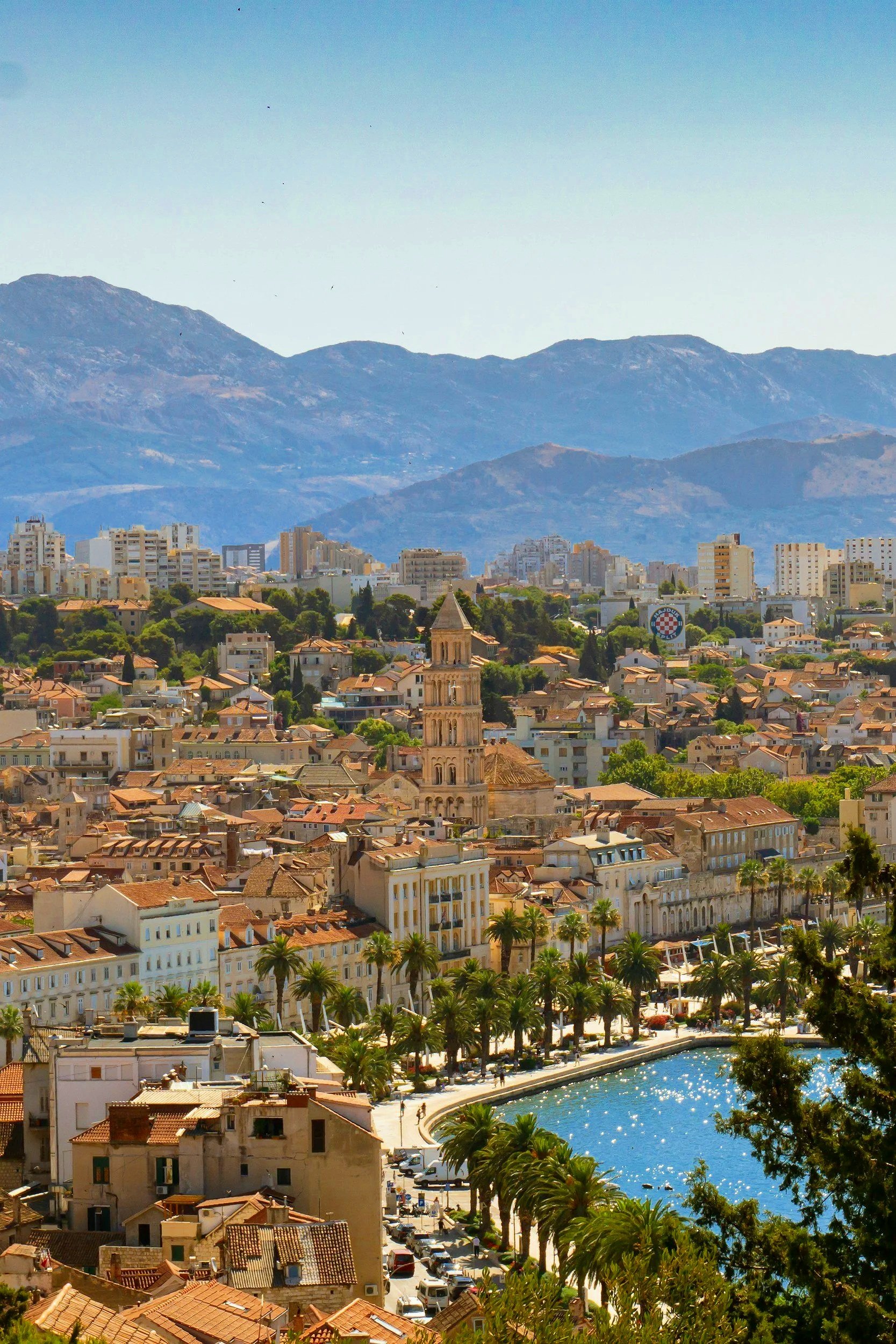 Vista de una ciudad costera con edificios históricos, palmeras y un lago, con montañas en el fondo.