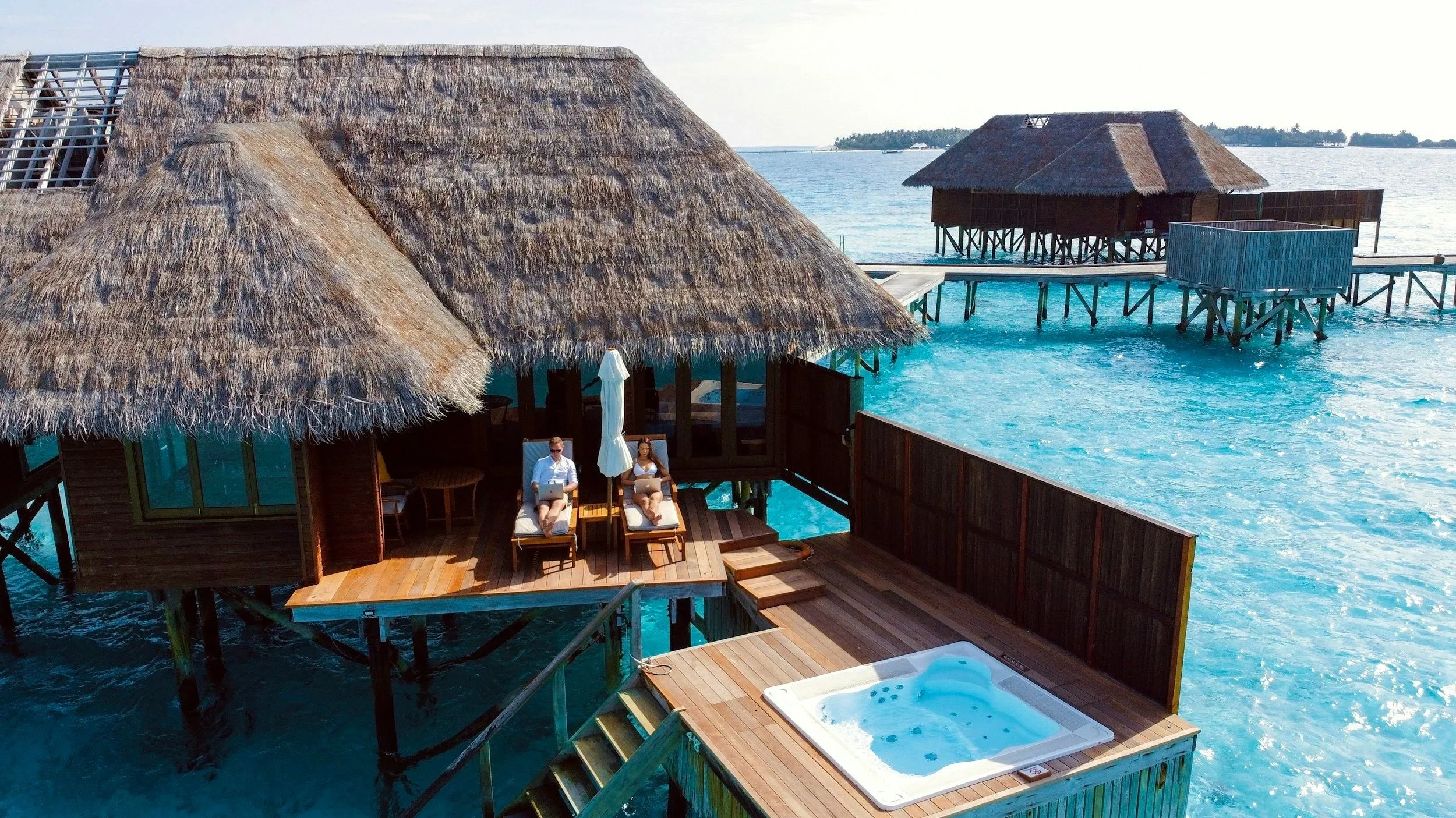 Cabañas en el agua con terraza, jacuzzi y muelles desde donde salen botes, en un ambiente tropical, con dos personas descansando en sillas lounge.