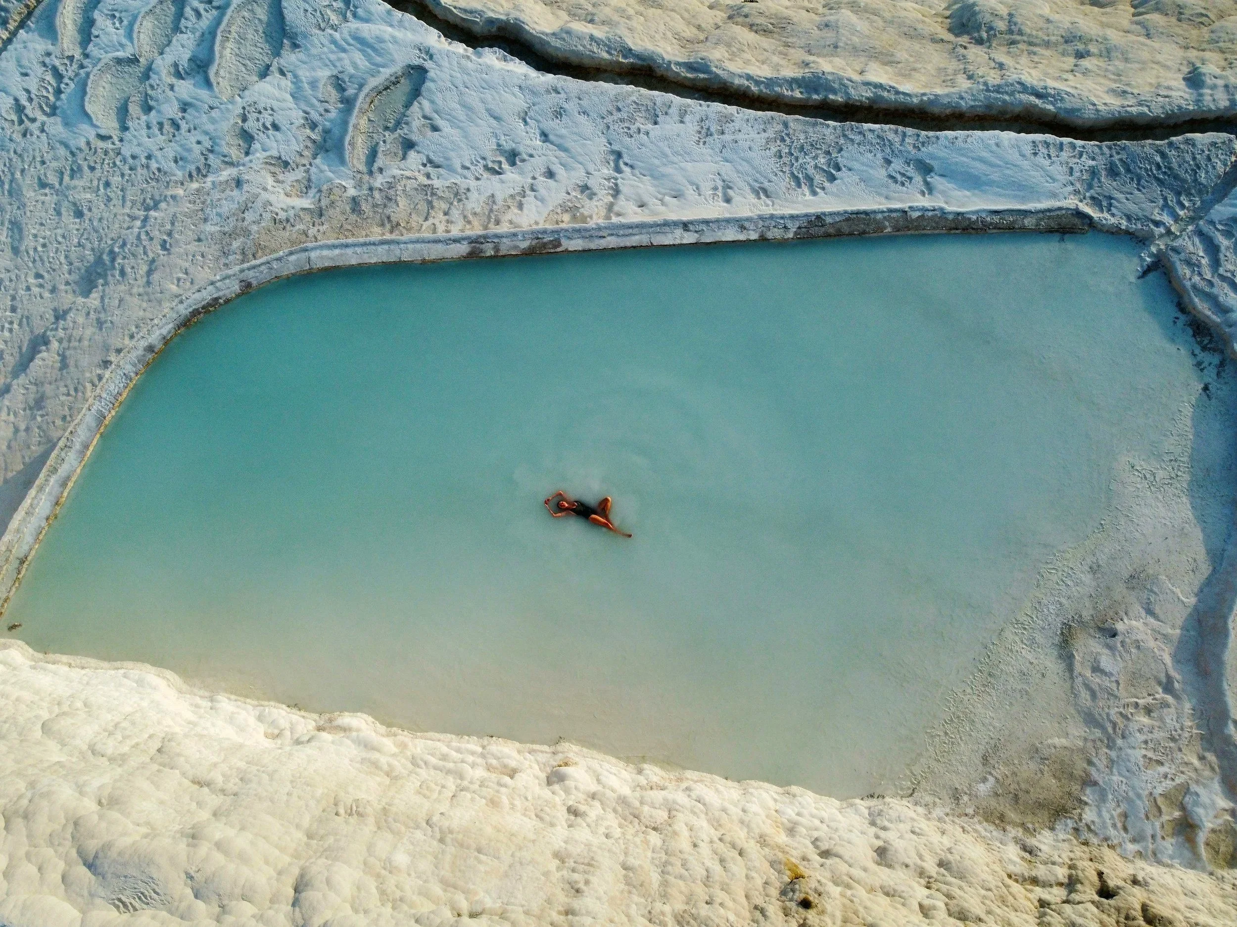 Persona flotando en una piscina de agua turquesa, rodeada por paredes blancas y piedras, en un ambiente seco y rocoso.