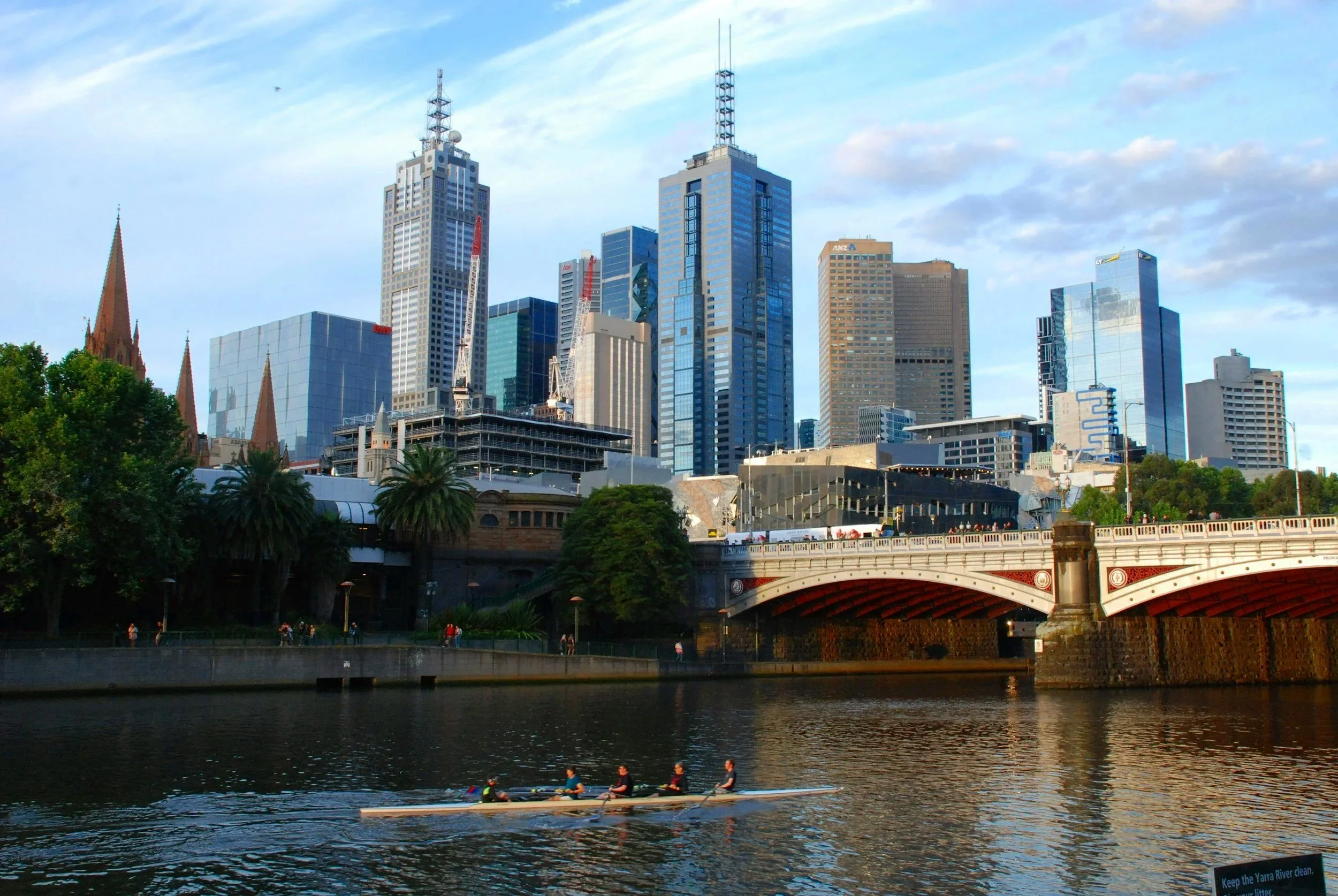 Vista de la ciudad con rascacielos y puente peatonal, reflejado en el río Yarra, con cinco personas en un bote remando.