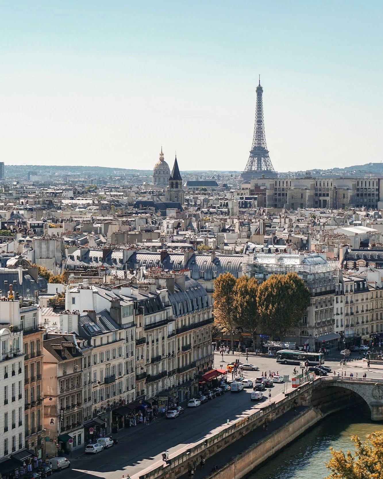 Vista panorámica de París con la Torre Eiffel y otros edificios históricos al fondo, río Sena y muelle con tráfico y peatones en primer plano.