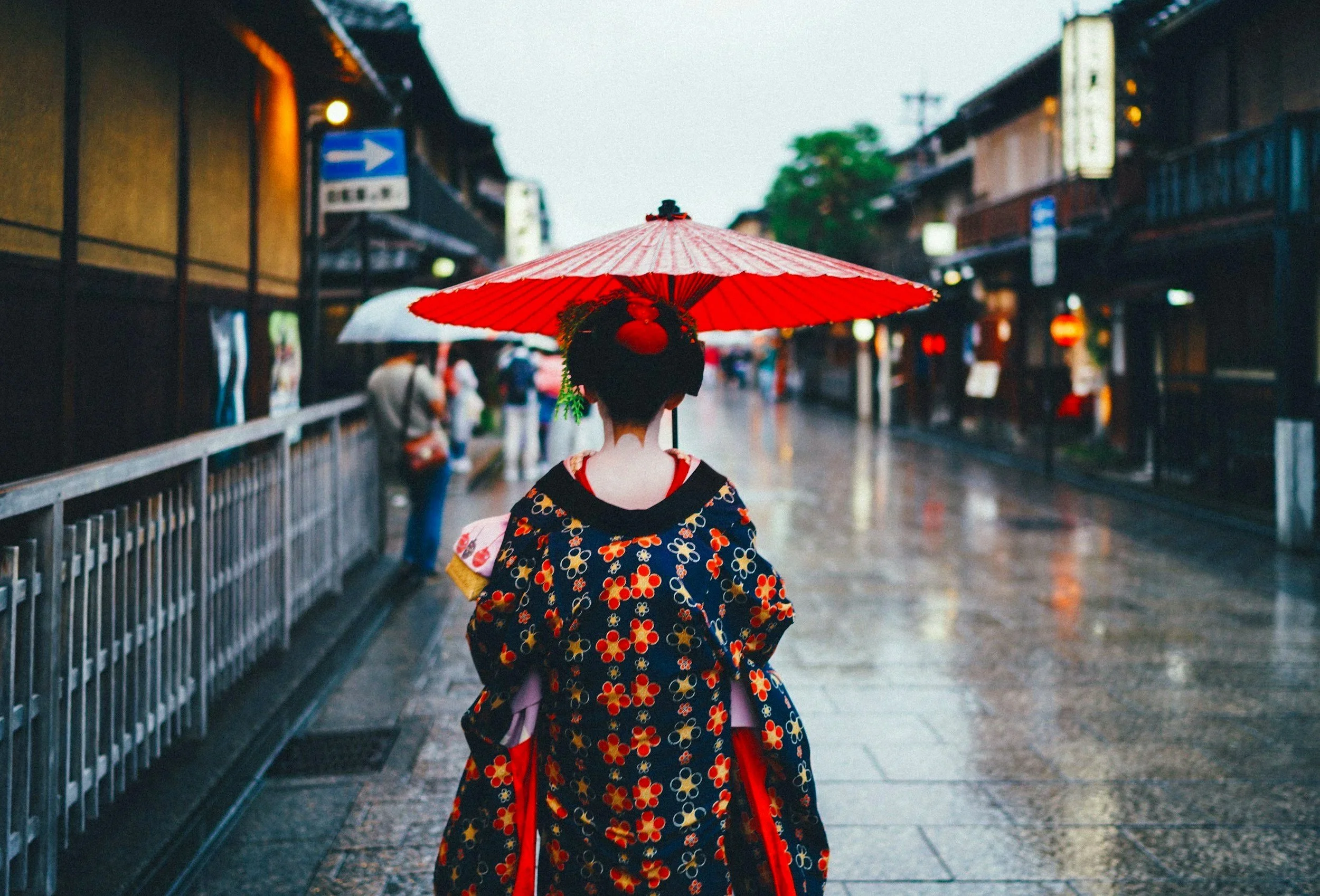 Mujer vestida con kimono tradicional japonés, con peinado elaborado, sosteniendo paraguas rojo en calle lluviosa con tiendas y personas en el fondo.
