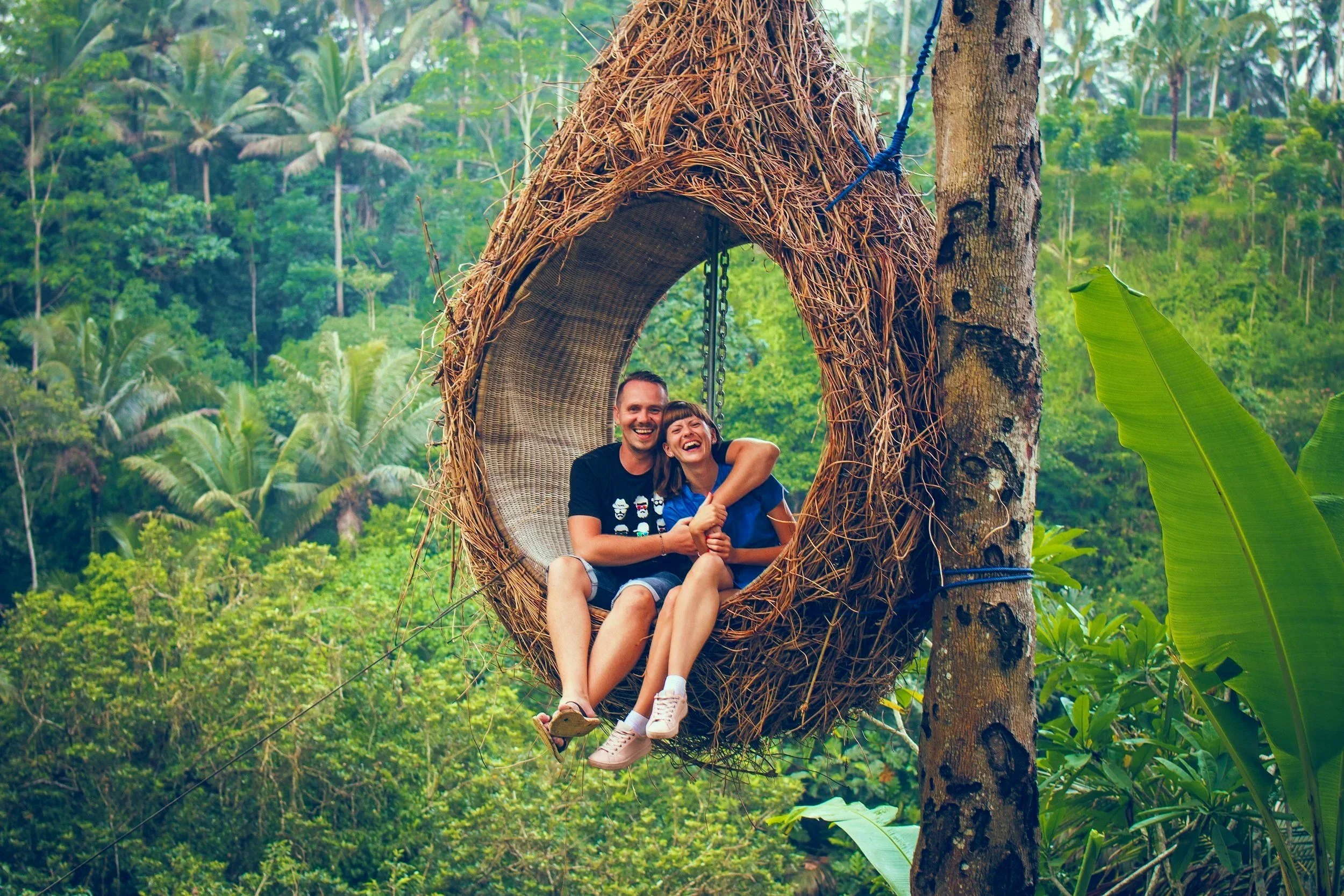 Pareja sonriendo y abrazada en una hamaca de mimbre colgada en un árbol en medio de una selva tropical con muchas plantas verdes.