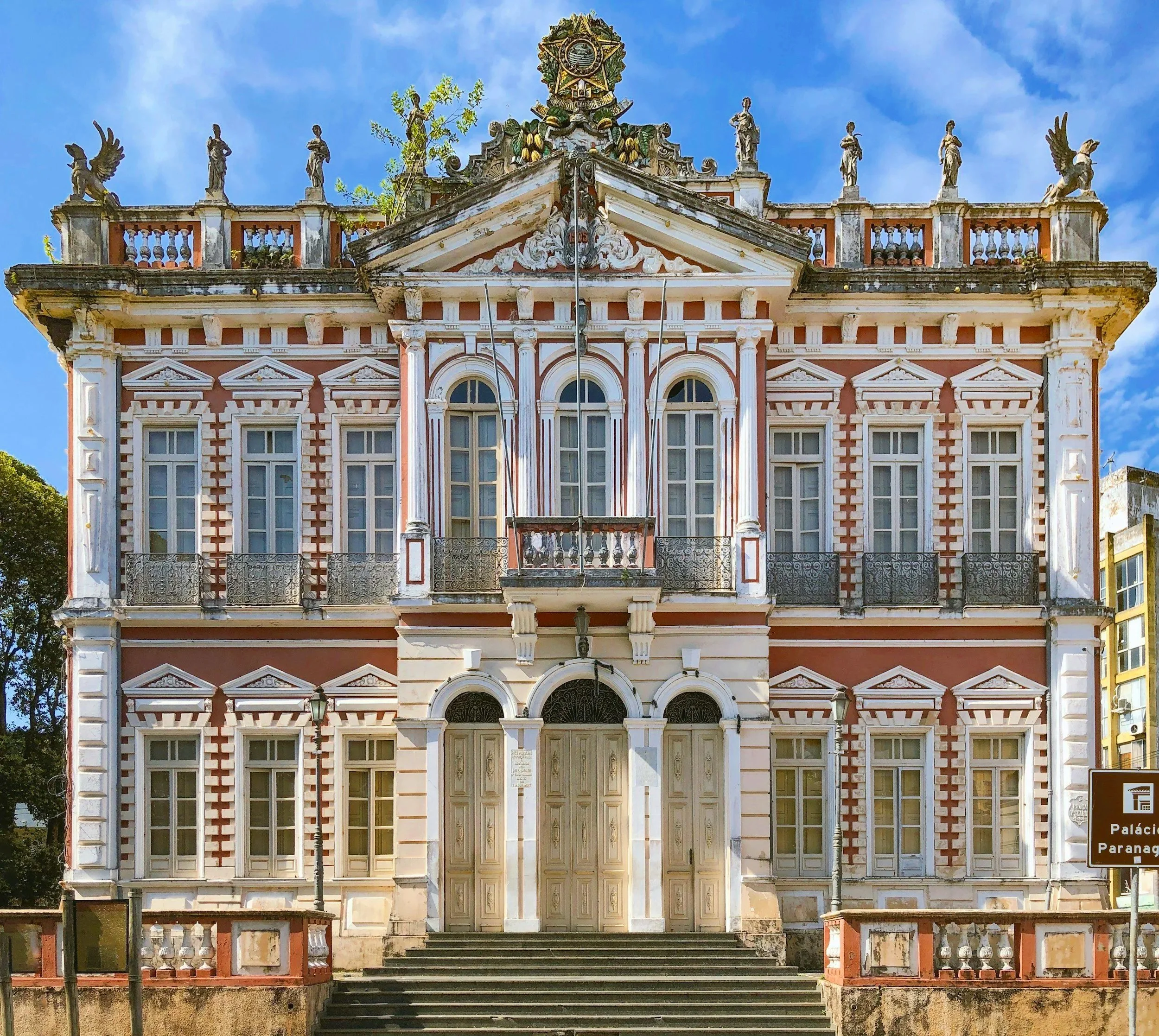 Edificio antiguo de estilo colonial con fachada rosada y detalles blancos, varias ventanas altas con marcos blancos, puerta principal de madera con arcos y escaleras, cartel que indica "Palacio Paranágica" y escultura en la azotea.
