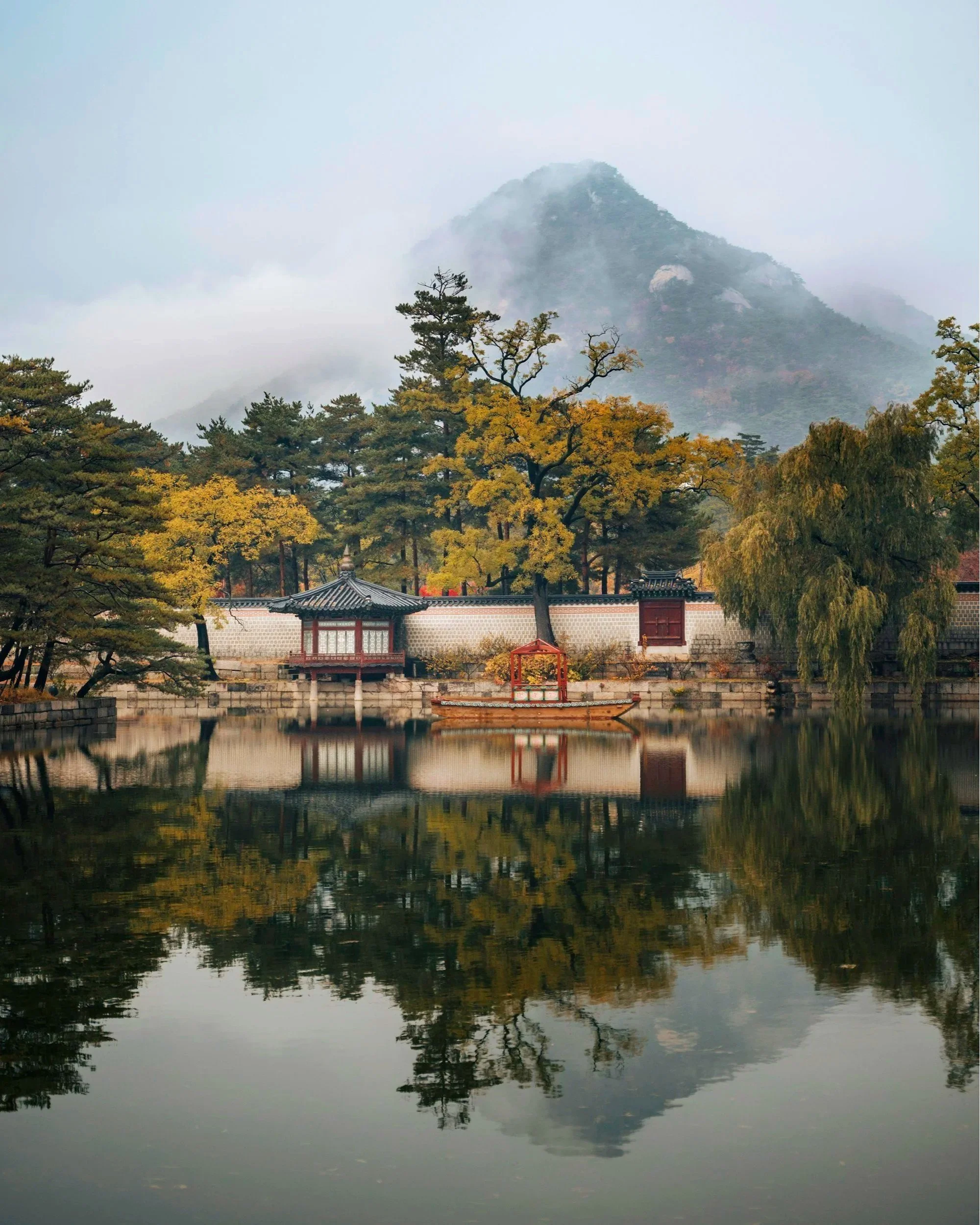 Paisaje de un lago con árboles otoñales, un pabellón tradicional y un bote, con una montaña envuelta en niebla de fondo.