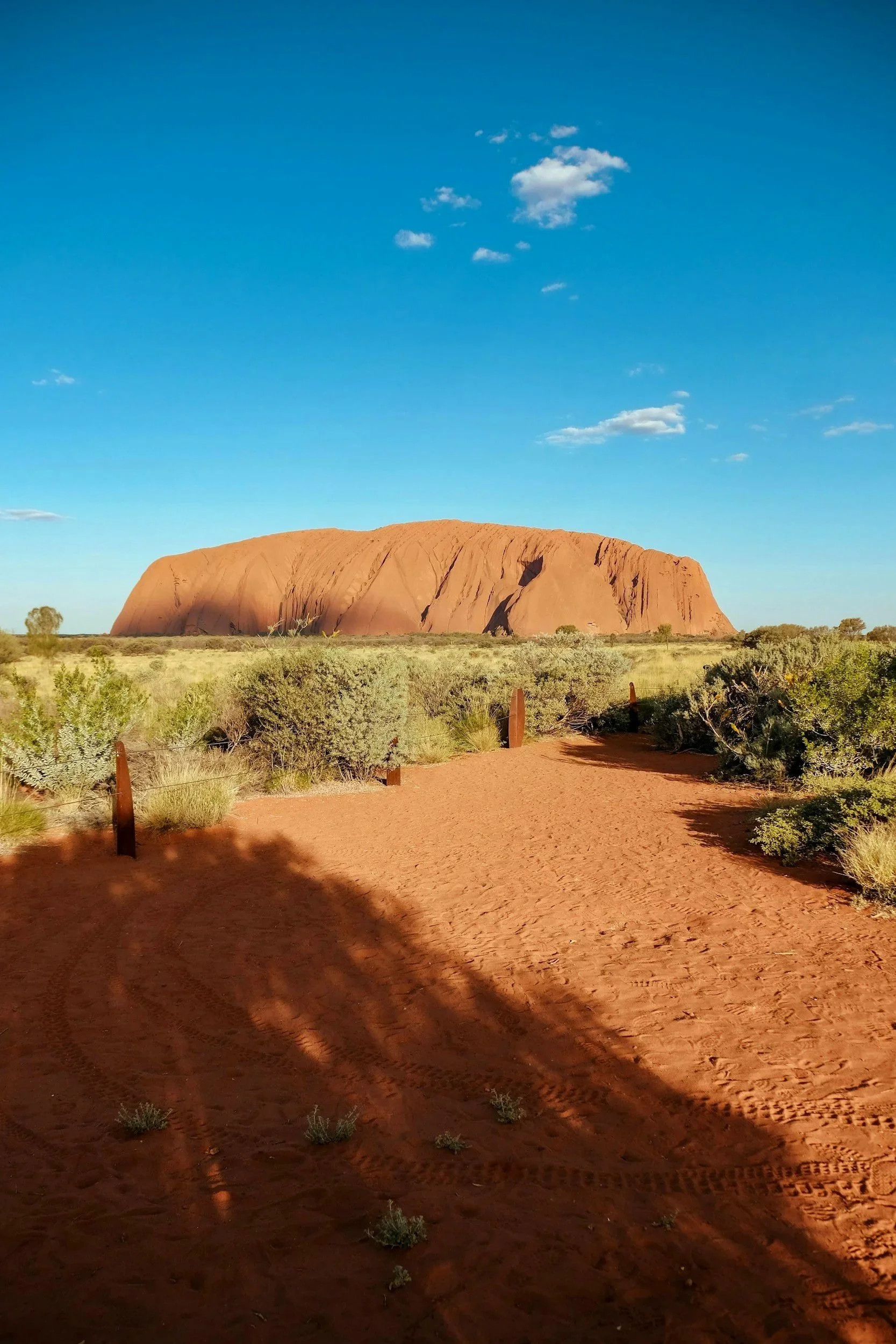 Paisaje desértico con la roca Uluru en el fondo, bajo un cielo azul con algunas nubes, vegetación escasa y camino de arena en primer plano.