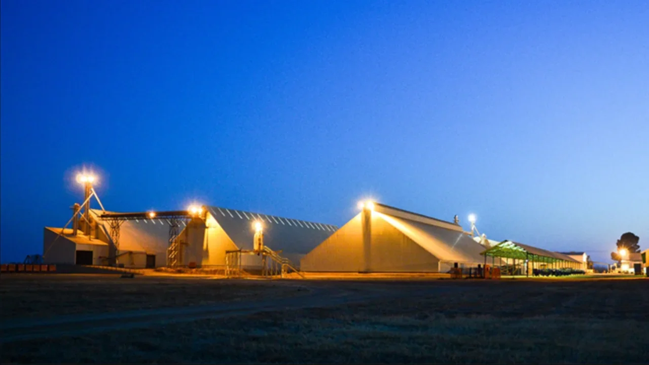 A series of illuminated barn-like structures at dusk or night with a clear blue sky and open area in the foreground.