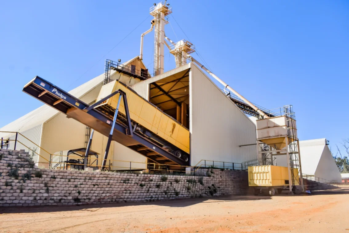 Industrial mining or processing facility with large metal structures, conveyor belts, and equipment against a blue sky.