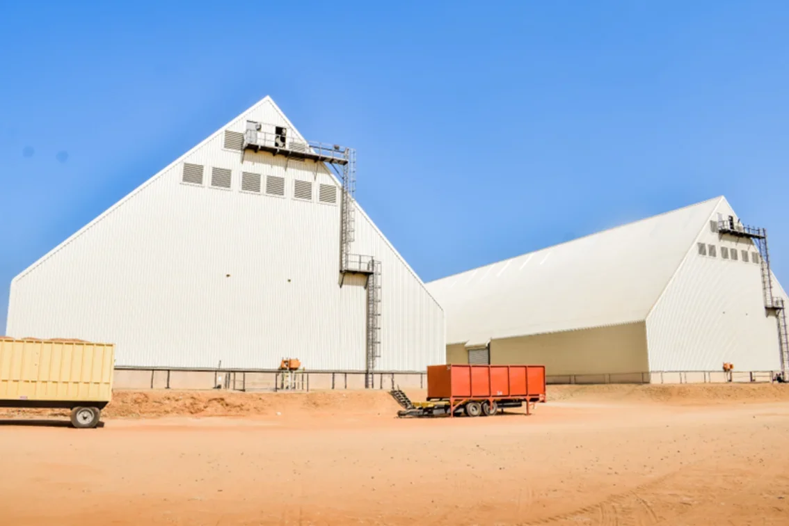 Two large white industrial buildings with sloped roofs under a clear blue sky. There are shipping containers and equipment on the ground in front of the buildings.
