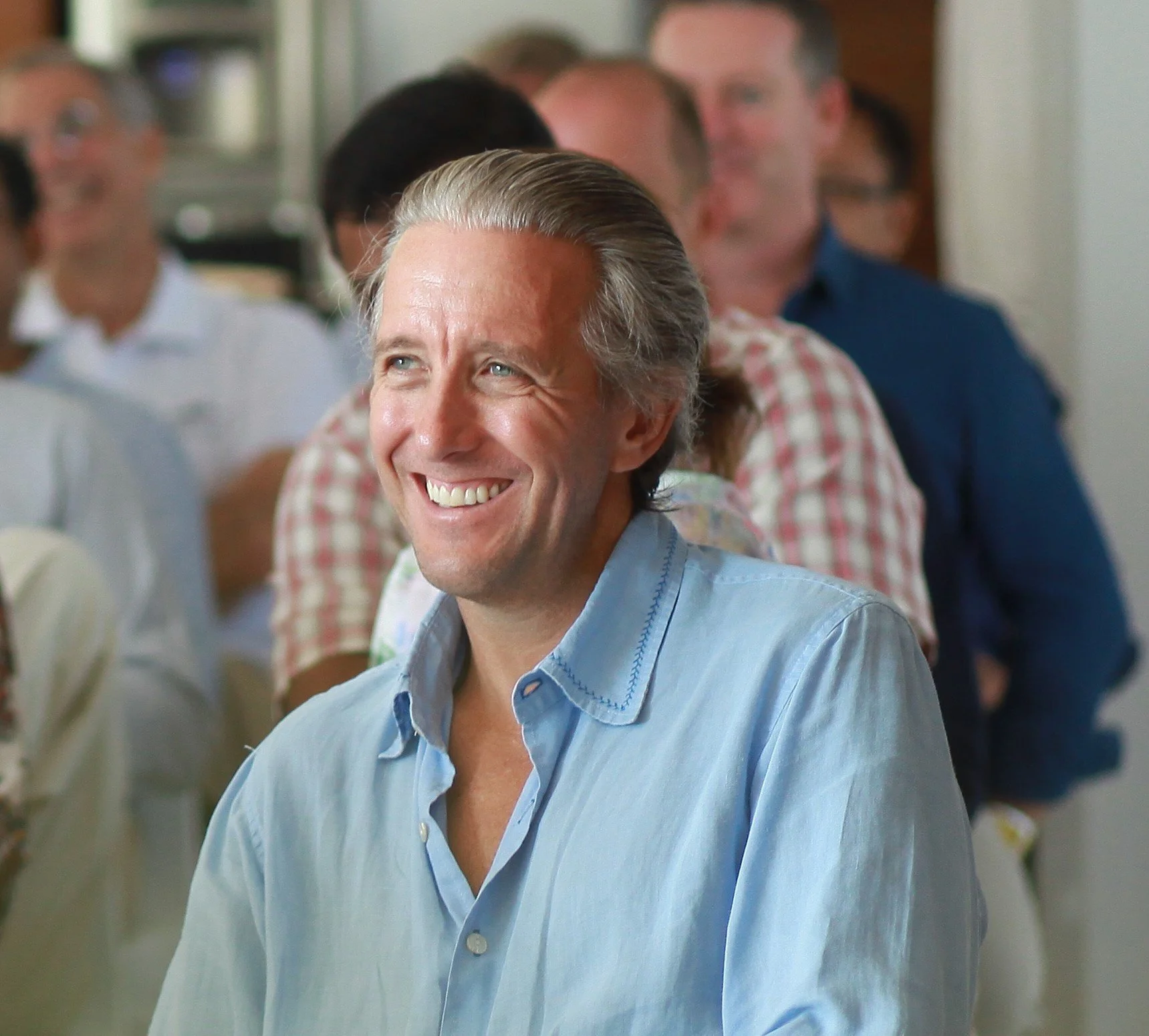 Smiling middle-aged man with gray hair in a light blue shirt at a social gathering with other people in the background.