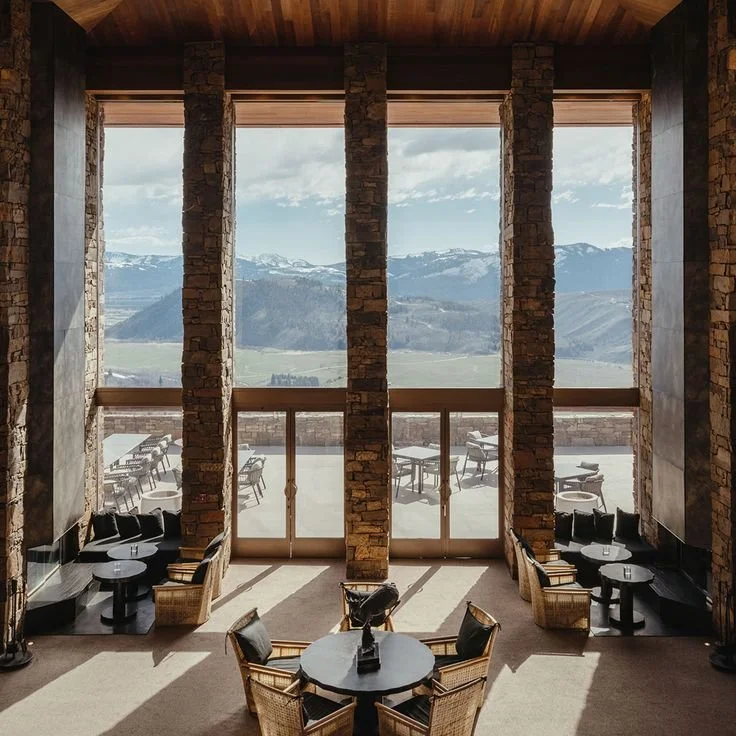 Interior view of a spacious lobby lounge area of Amangani hotel with large windows showing the Grand Tetons. The room features seating with tables, some with chairs and some with built-in banquettes around two open fireplaces, and a wooden ceiling.