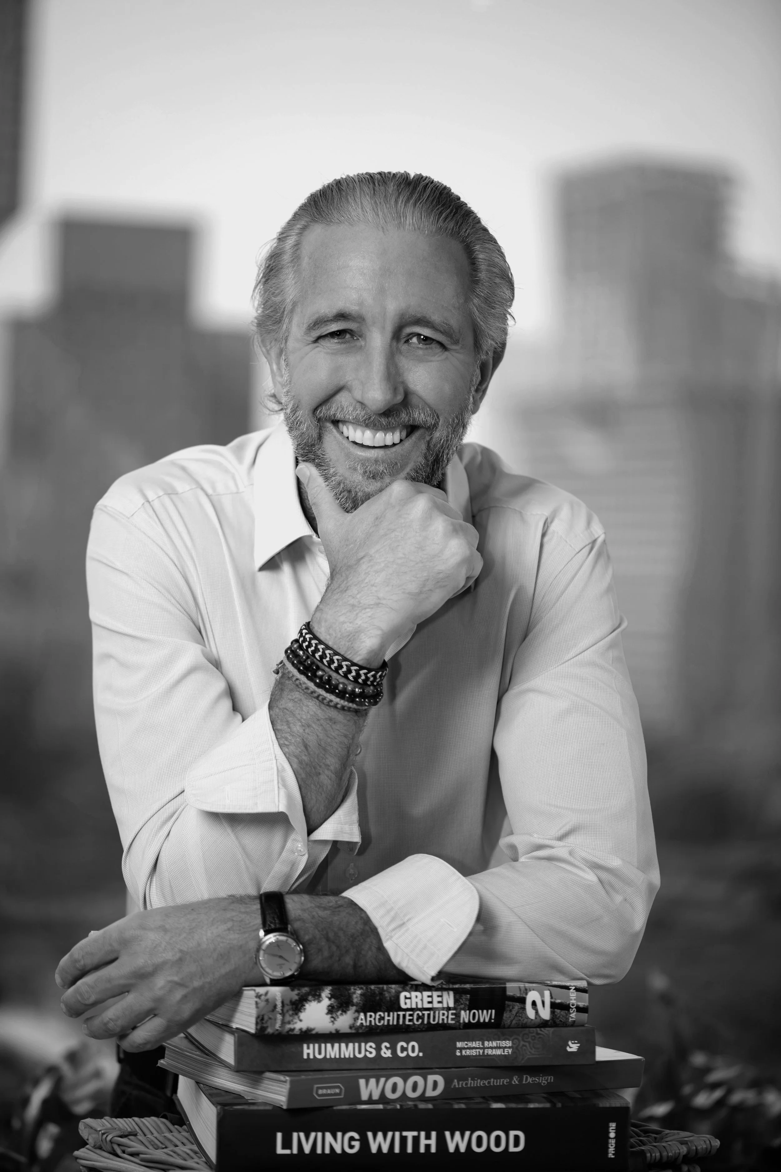 A smiling middle-aged man, Guy Heywood, with a beard and mustache, wearing a white dress shirt, glasses, and bracelets, is seated outdoors with a blurred cityscape in the background. He has a stack of books on architecture and design in front of him.
