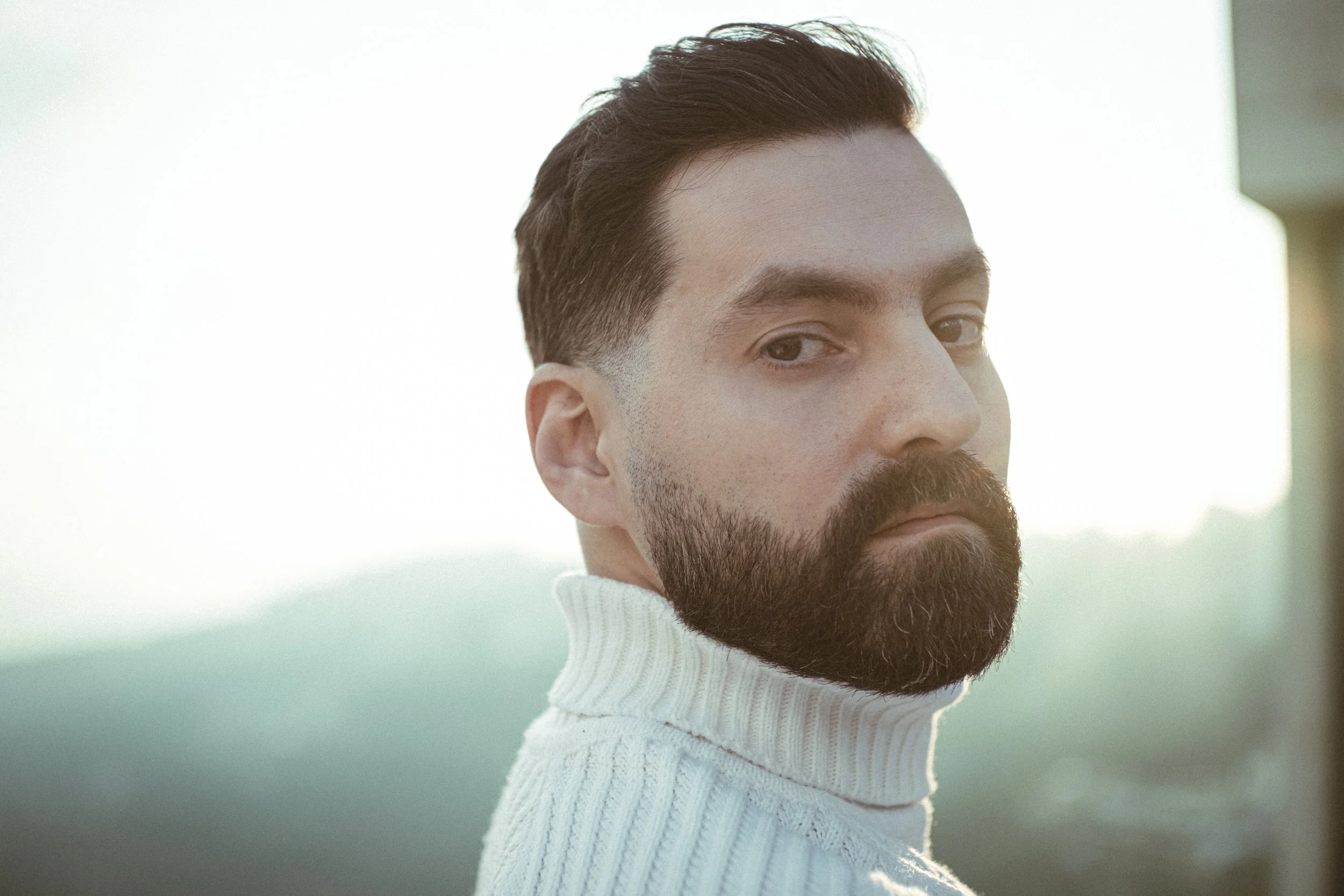 A man with dark hair and a beard looking over his shoulder while wearing a white turtleneck sweater against a blurred outdoor background.