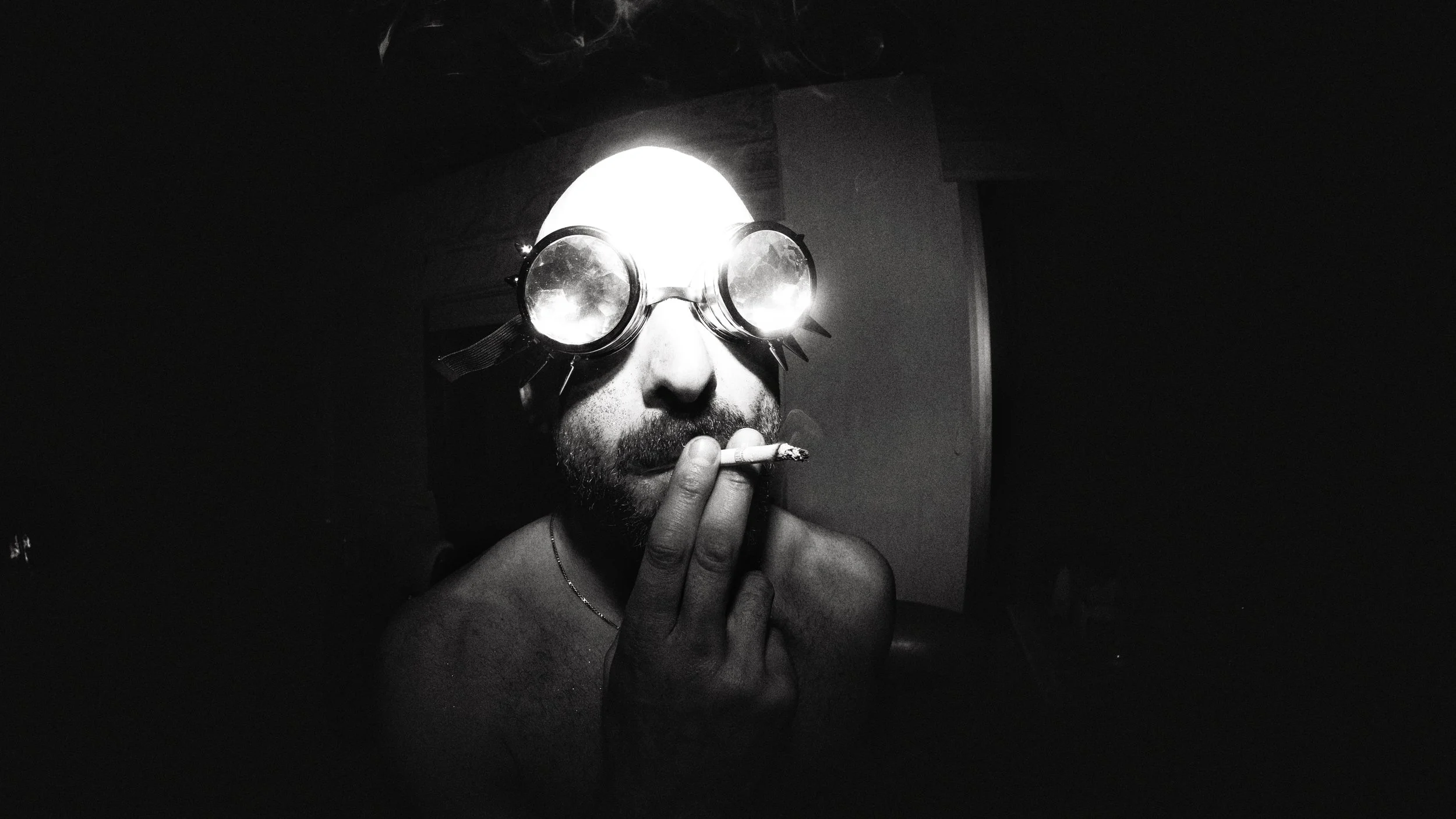 A shirtless man with a beard, wearing goggles and a headlamp, holding a cigarette near his mouth in a dark environment.