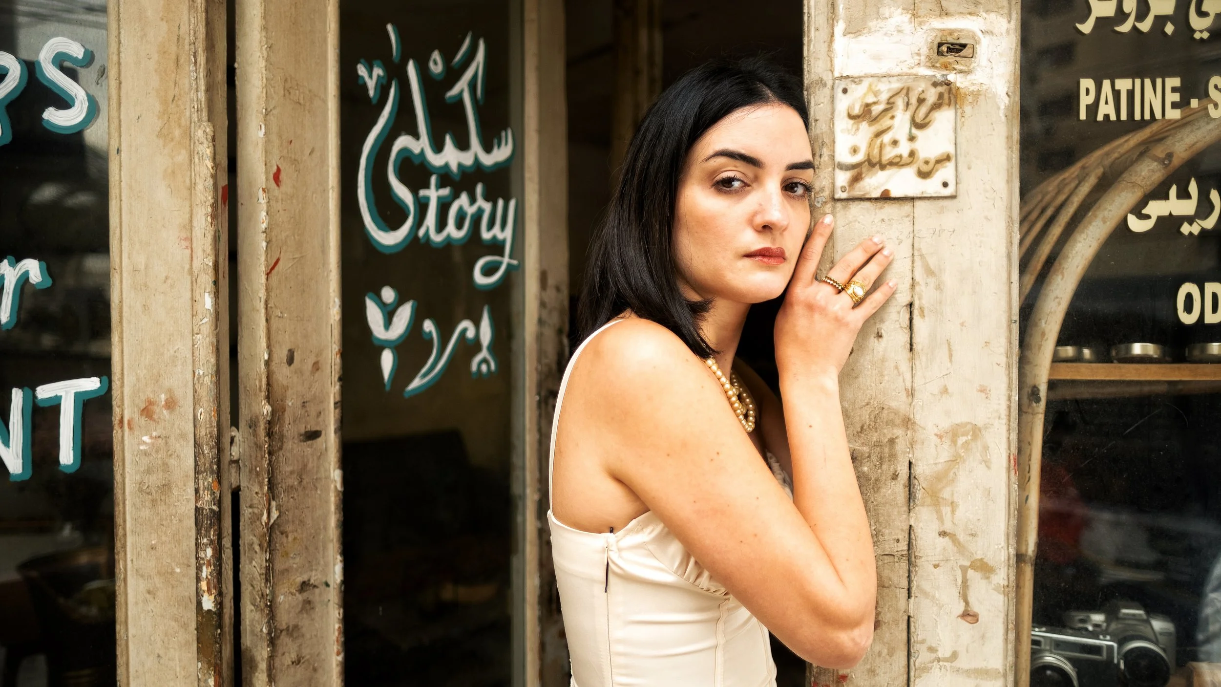 A woman with black hair and fair skin standing against a worn wall outside a shop, wearing a cream-colored satin dress and layered pearl necklaces, looking at the camera with a serious expression.