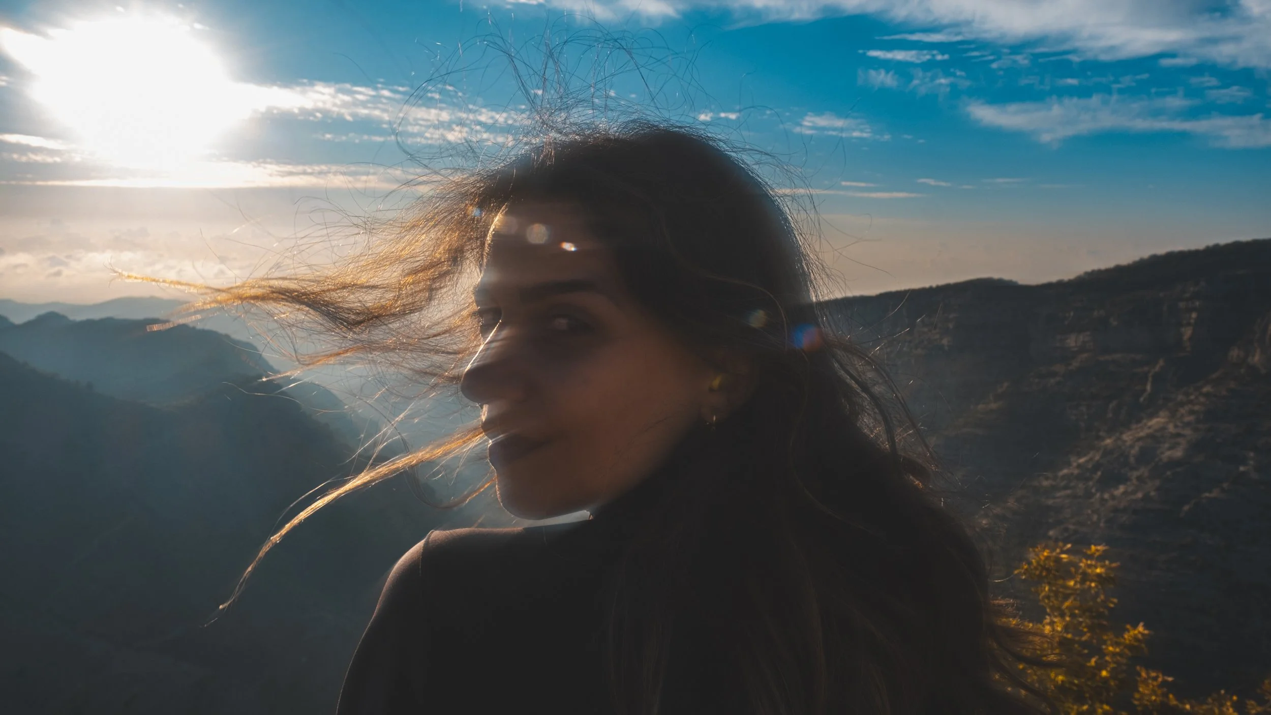 A woman with long hair blowing in the wind, standing against a mountainous landscape with a bright sun and blue sky.