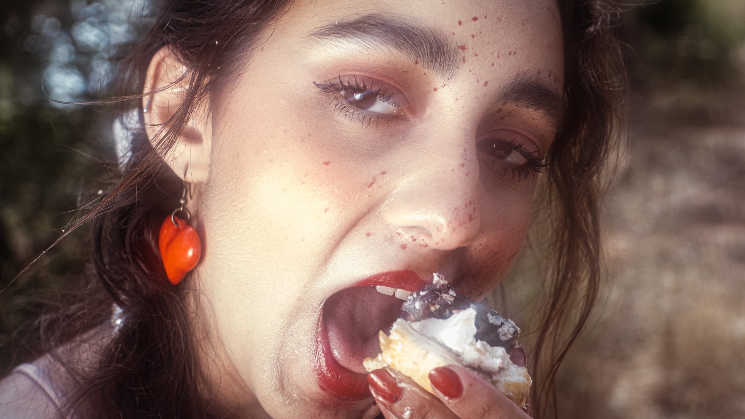 A woman with freckles and makeup, wearing red heart-shaped earrings, is eating a slice of cake with white frosting outdoors.
