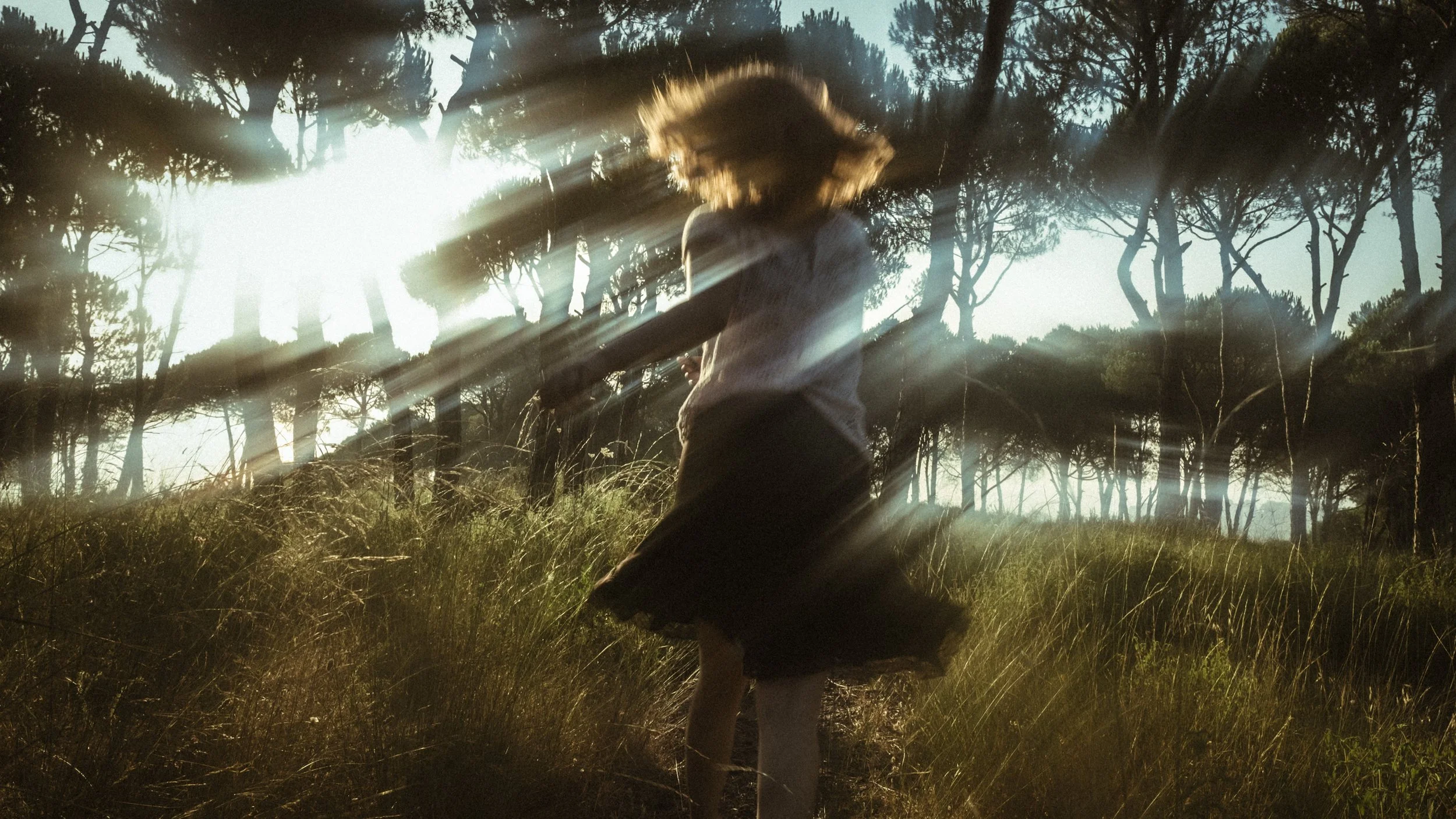 A person with shoulder-length hair wearing a skirt and blouse, standing in a grassy field surrounded by trees with sunlight streaming through, creating lens flare.