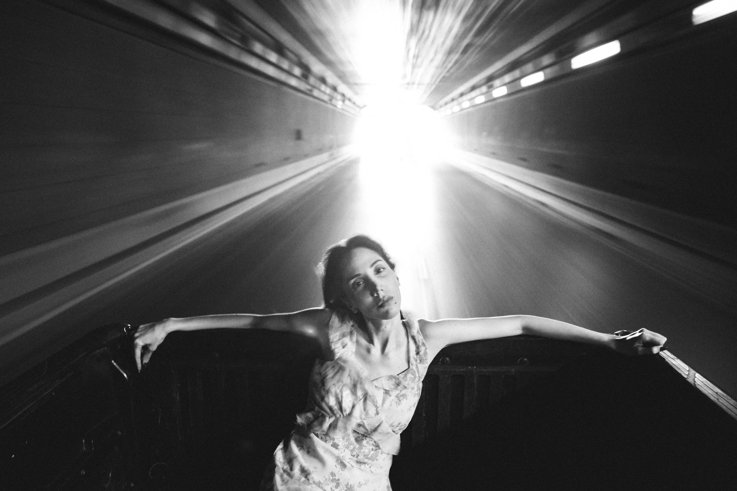 Black and white photo of a woman with arms outstretched, standing on a moving train with a bright tunnel and light at the end in the background.