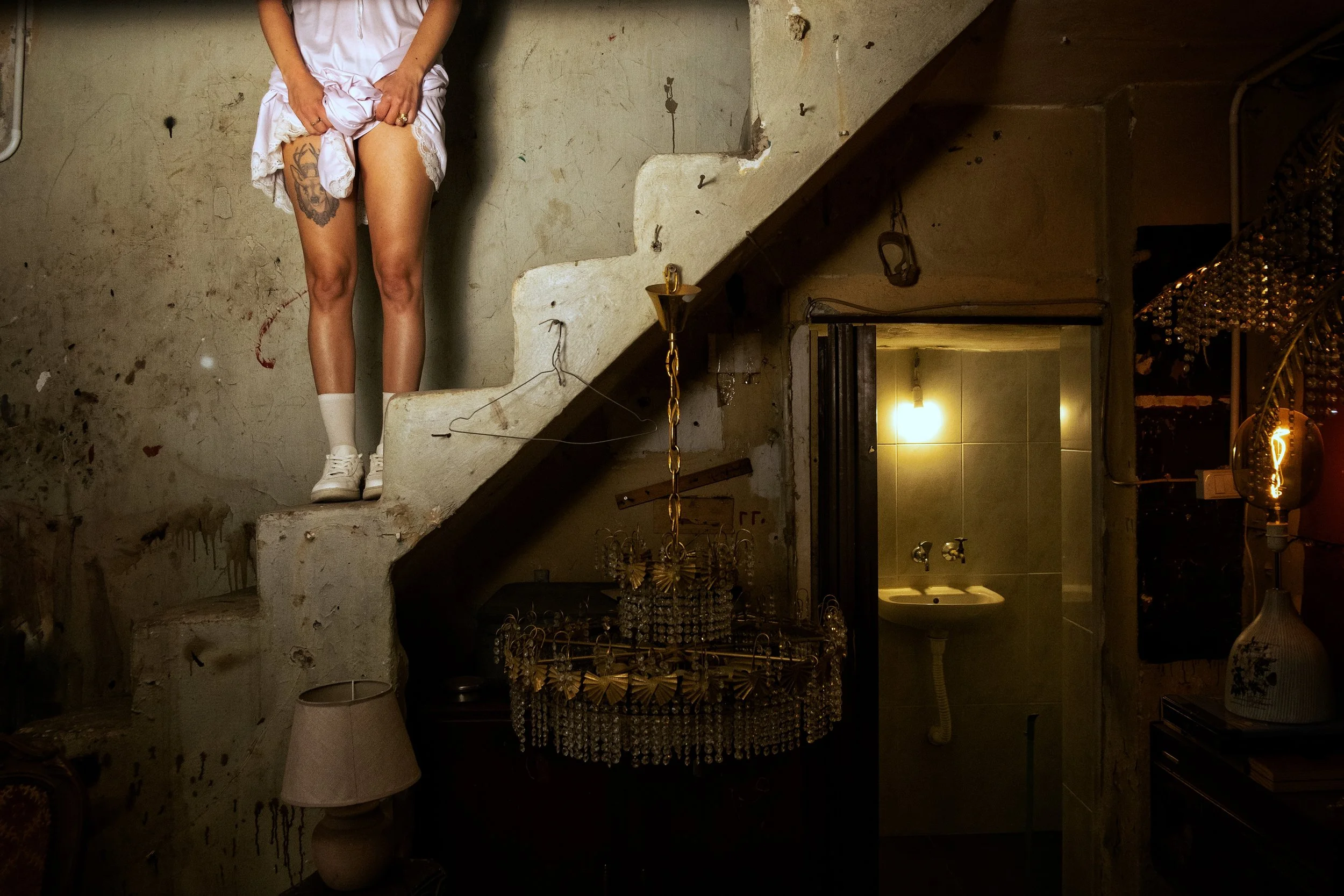 A person standing on a staircase, lifting a white dress to reveal their thigh tattoo. The setting appears to be an old, dimly lit room with a chandelier hanging from the ceiling and a rustic decor.