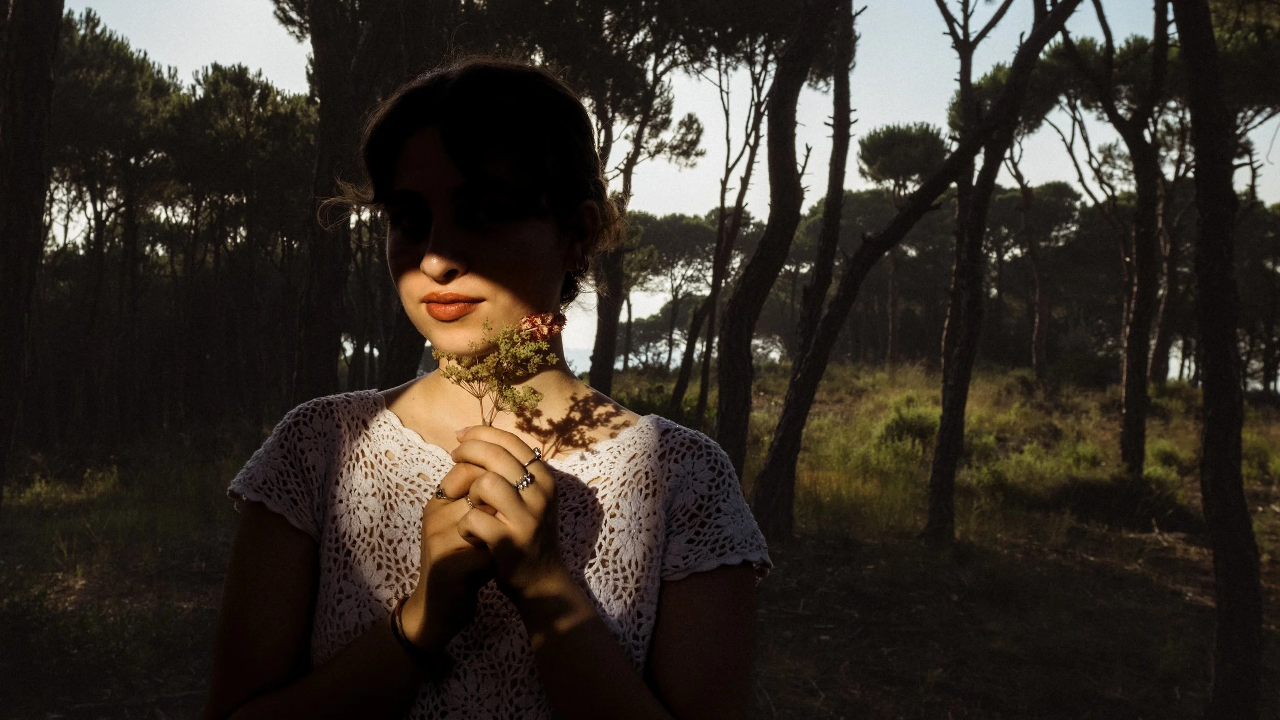 A woman holding a small bouquet of wildflowers in a forested area during sunset or sunrise.