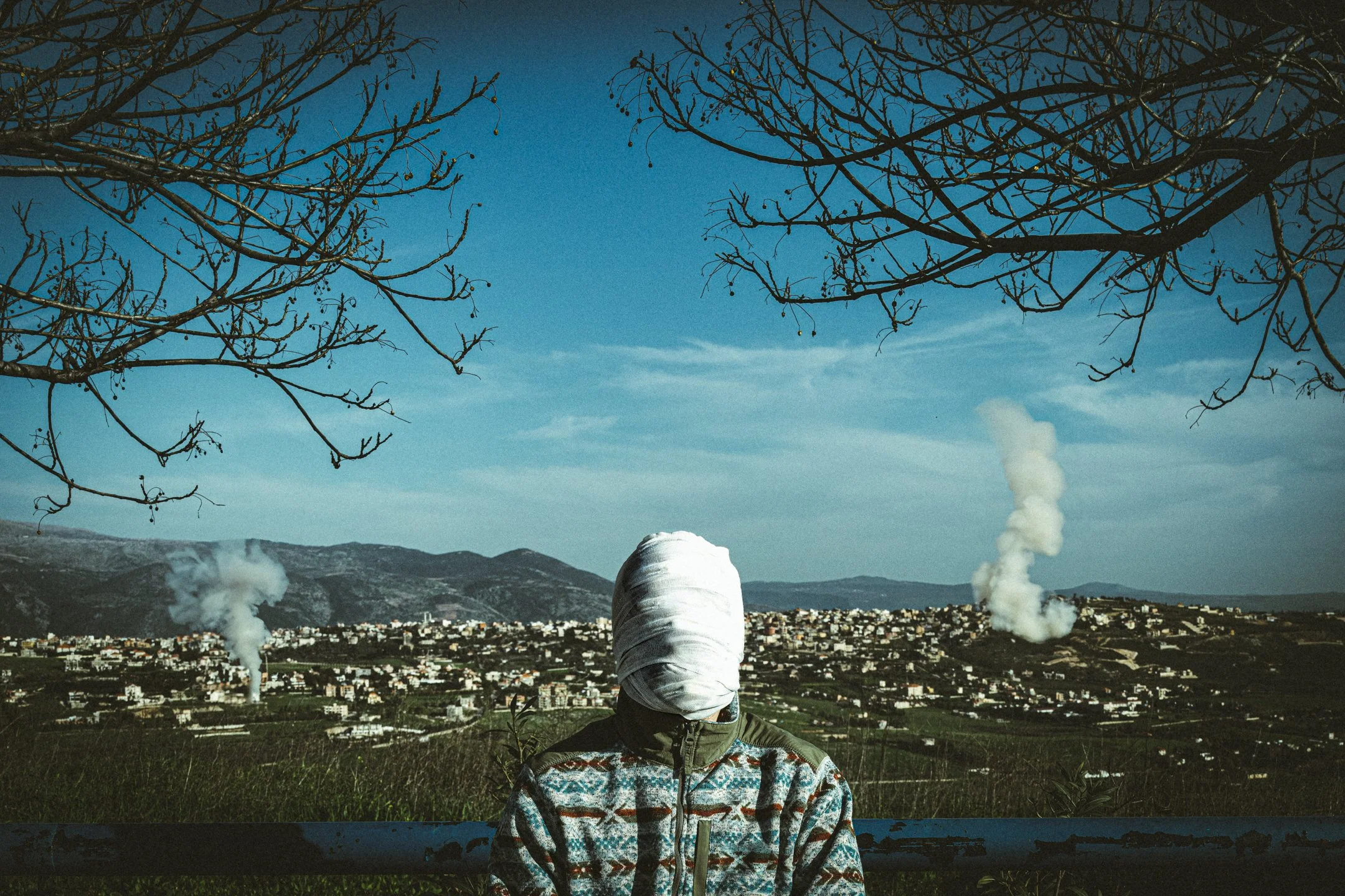 Person with a white cloth covering their head, sitting on a bench and overlooking a cityscape with industrial smoke emissions, hills, and blue sky with scattered clouds.