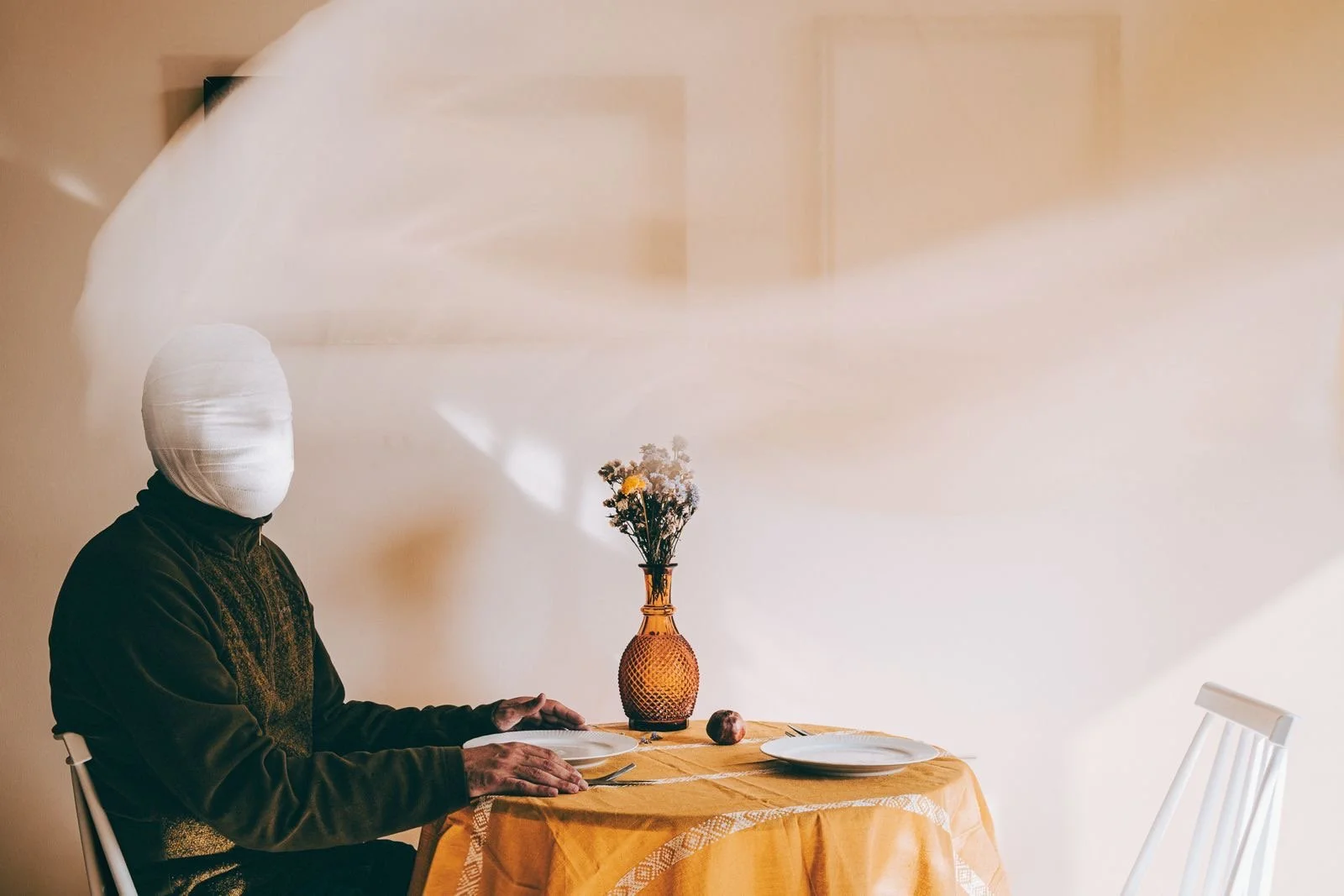 Person with face wrapped in white cloth sitting at a dining table with yellow tablecloth, empty plates, a fork and knife, a vase with flowers, and a red apple, against a plain beige wall.