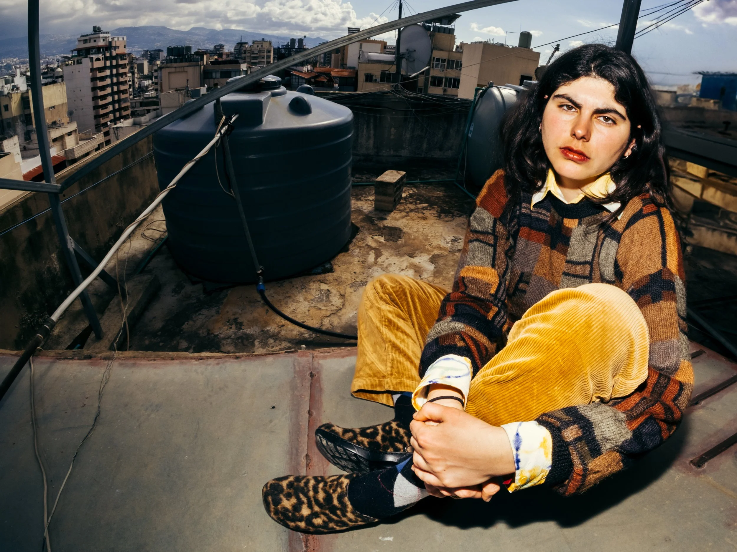 Young woman sitting on a rooftop in the city, wearing a patchwork sweater, mustard corduroy pants, leopard print shoes, and a collared shirt, with city buildings and mountains in the background.