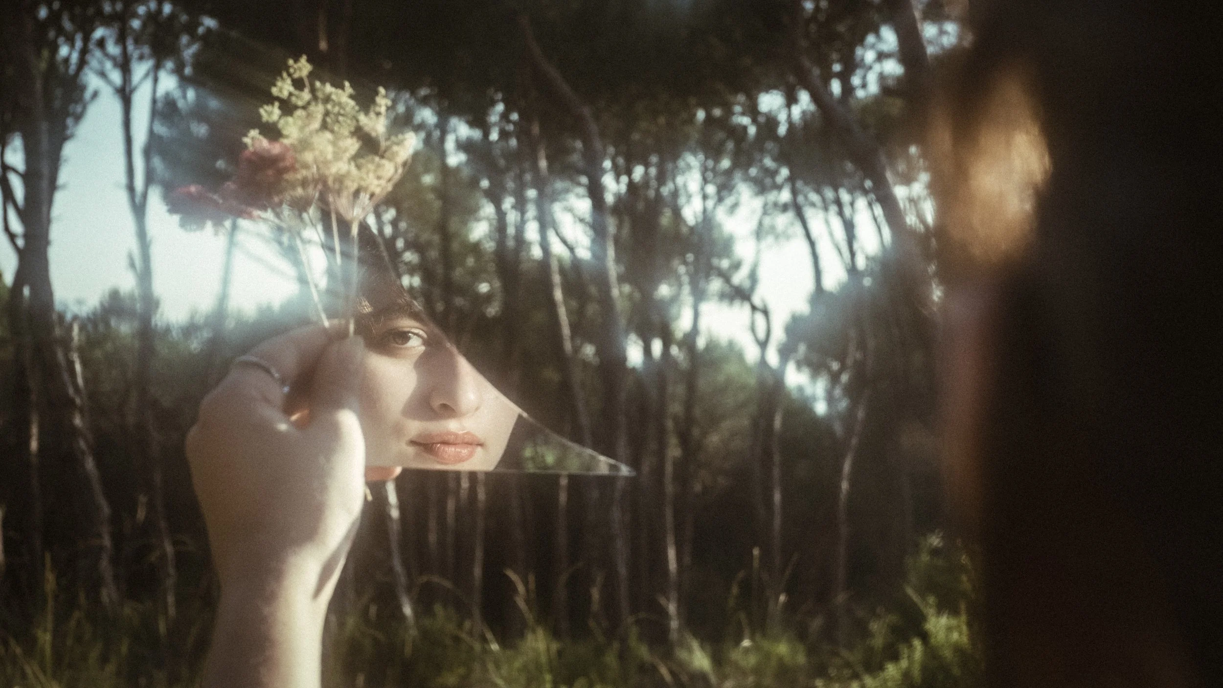 A person holding a broken mirror reflecting a young woman's face against a wooded outdoor background.