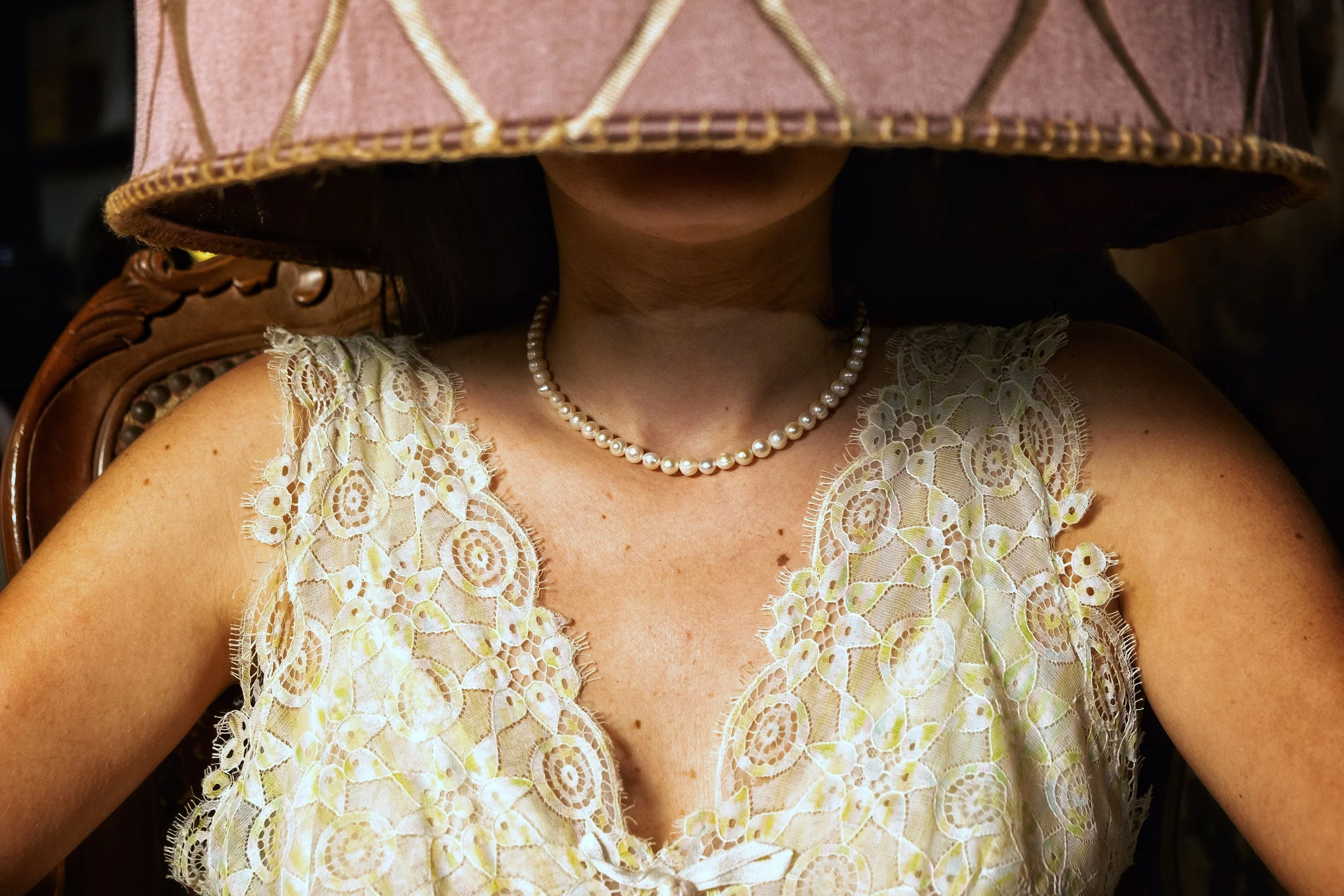 A woman wearing a large pink hat, a pearl necklace, and a lacy cream-colored dress sitting on a wooden chair.
