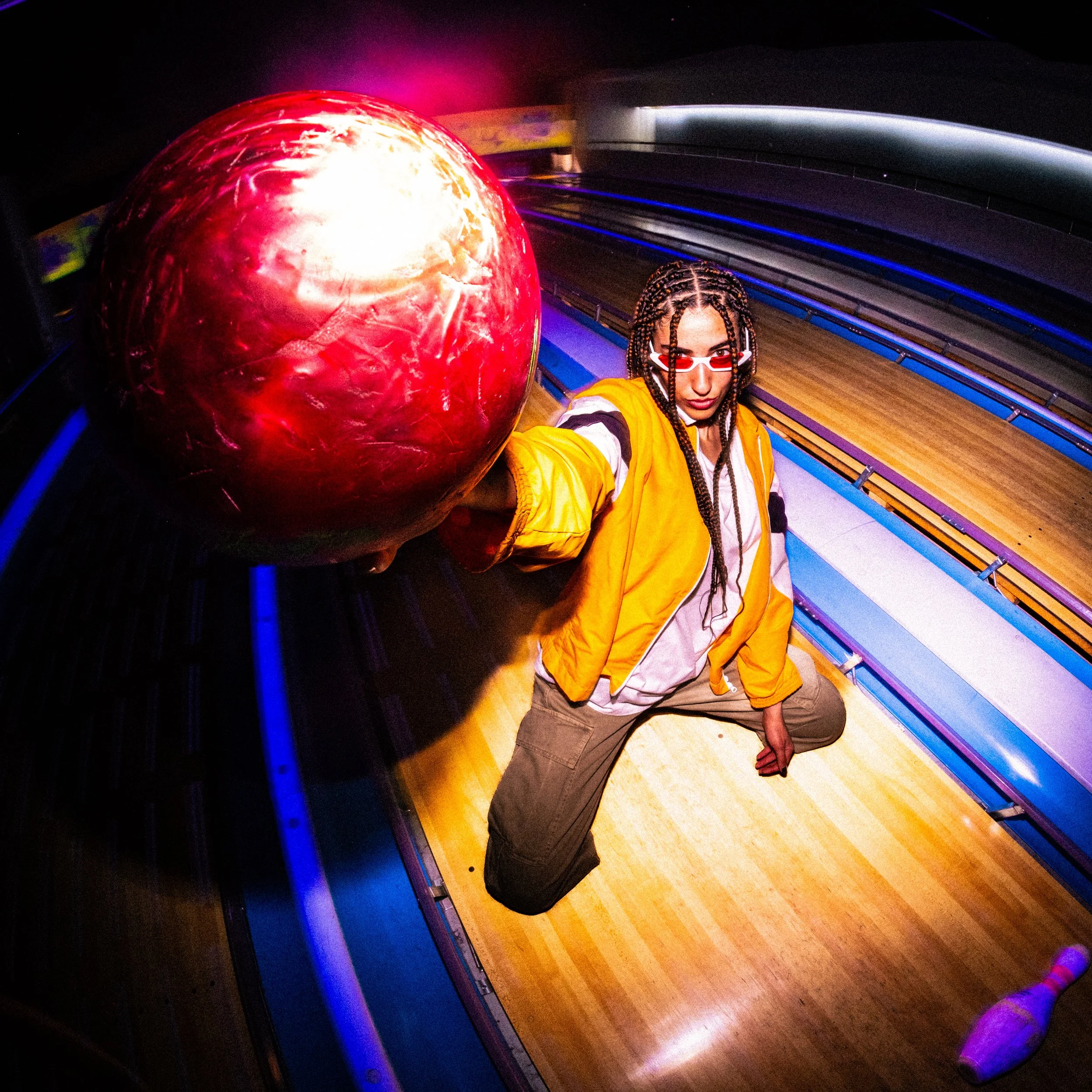 A woman with braided hair and red sunglasses kneels on the floor of a bowling alley, holding a large red bowling ball.
