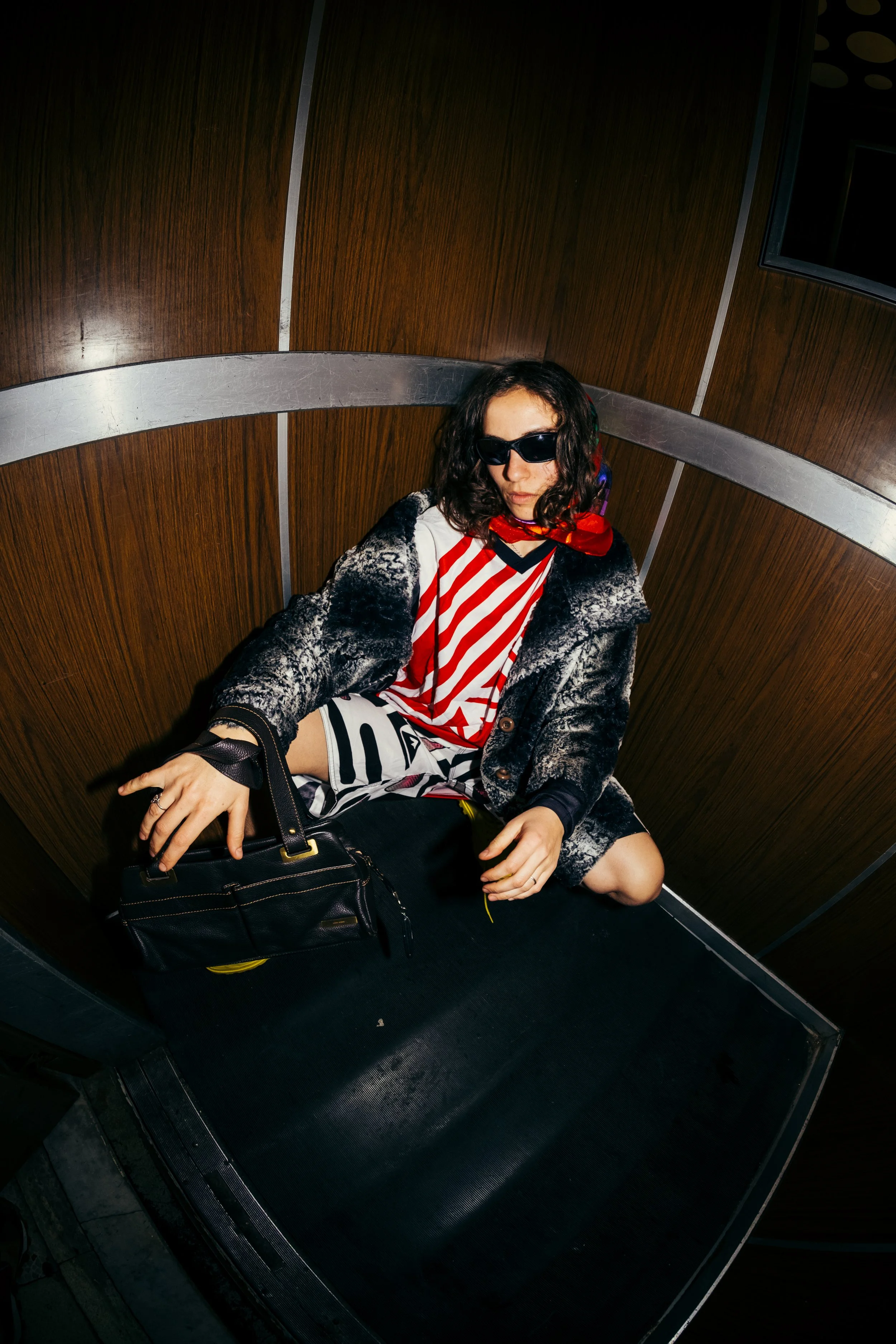 Person with curly hair wearing sunglasses, a red and white striped shirt, and a dark patterned jacket, sitting on an elevator floor with a black purse beside them.