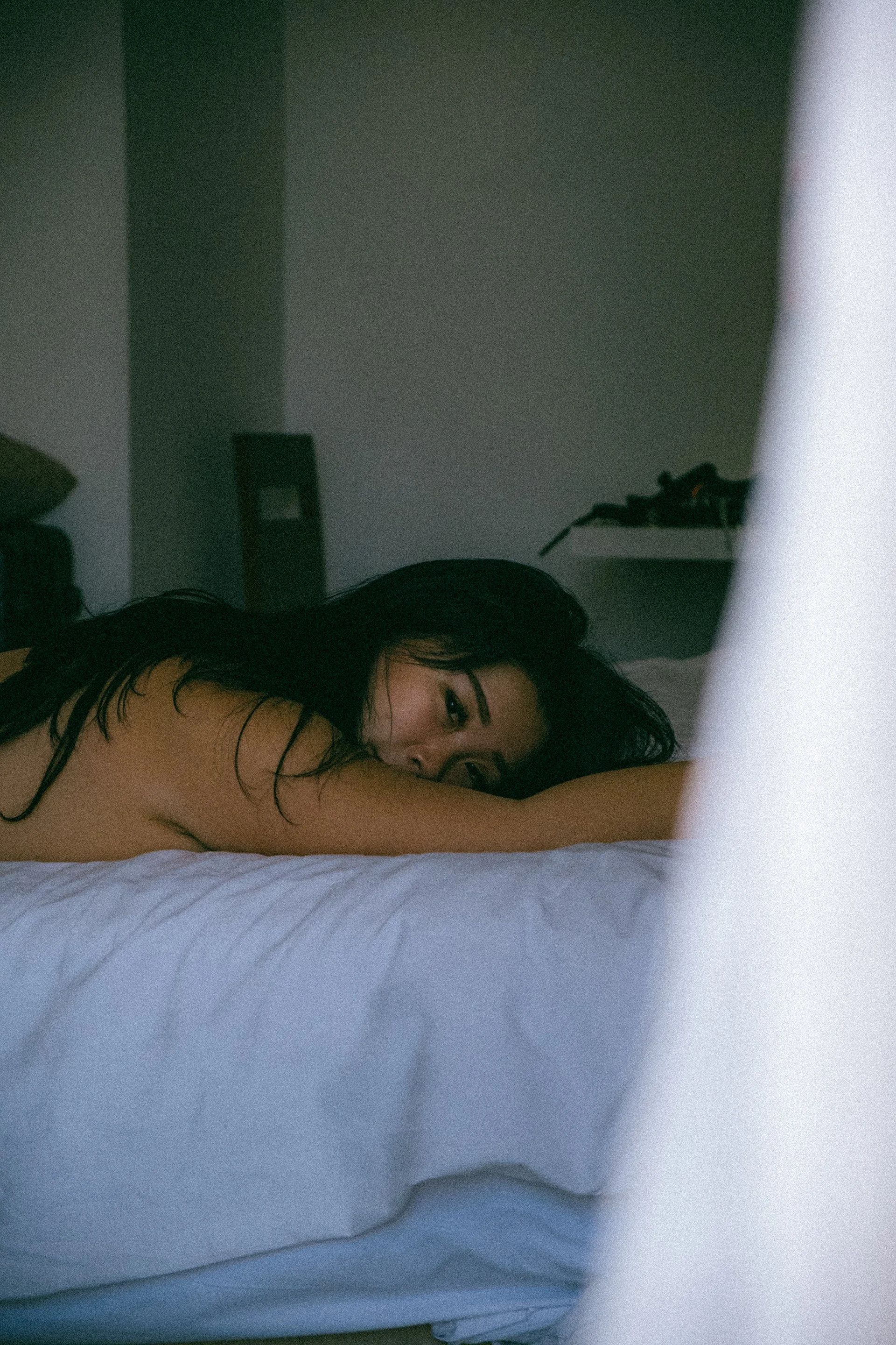 A woman with long black hair lying on her side on a bed, resting her head on her arm, with a partly visible white sheet and a dark wall in the background.