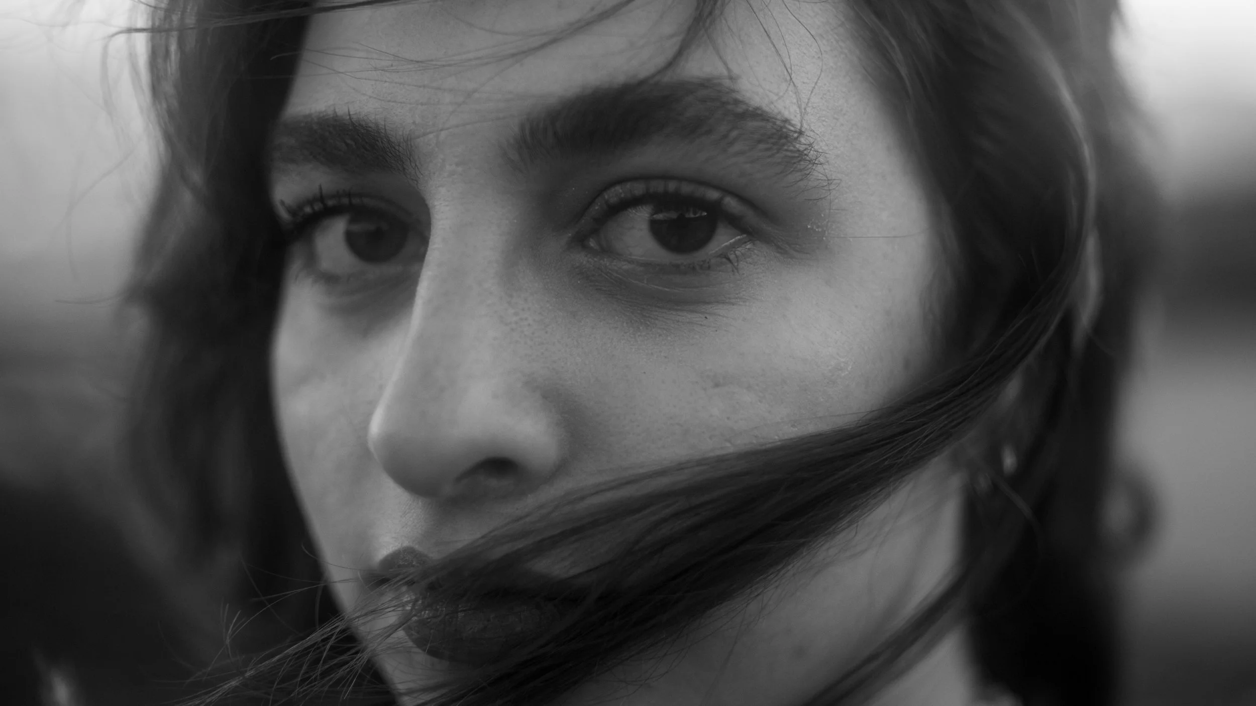 Close-up black and white photo of a woman's face with hair partially covering it, focusing on her eyes.