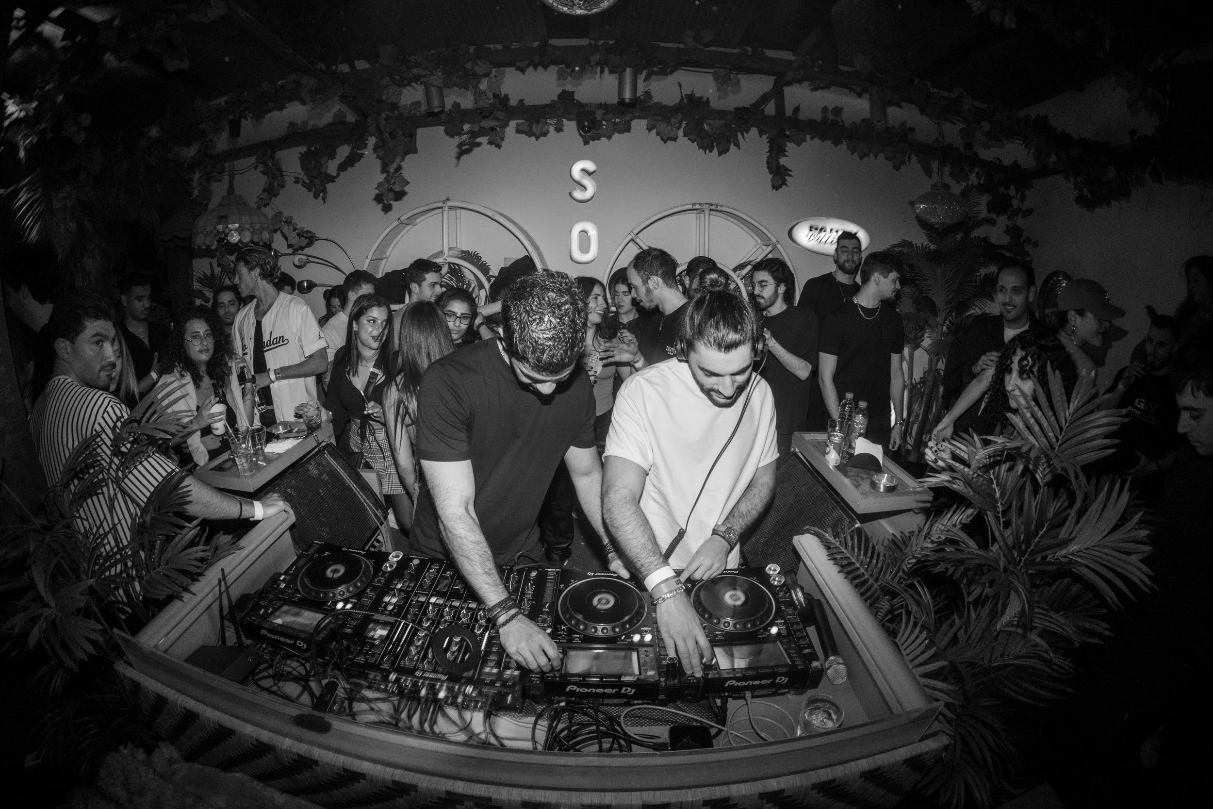 Two DJs mixing music at a party with a crowd of young people dancing and socializing in a decorated indoor venue.