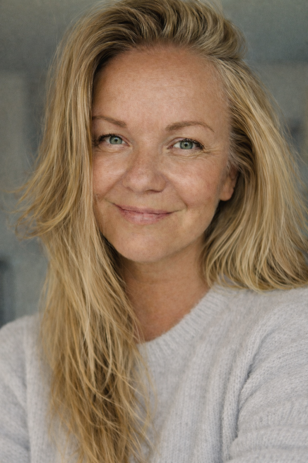 Close-up of a smiling woman with long blonde hair and blue eyes, wearing a light-colored sweater.