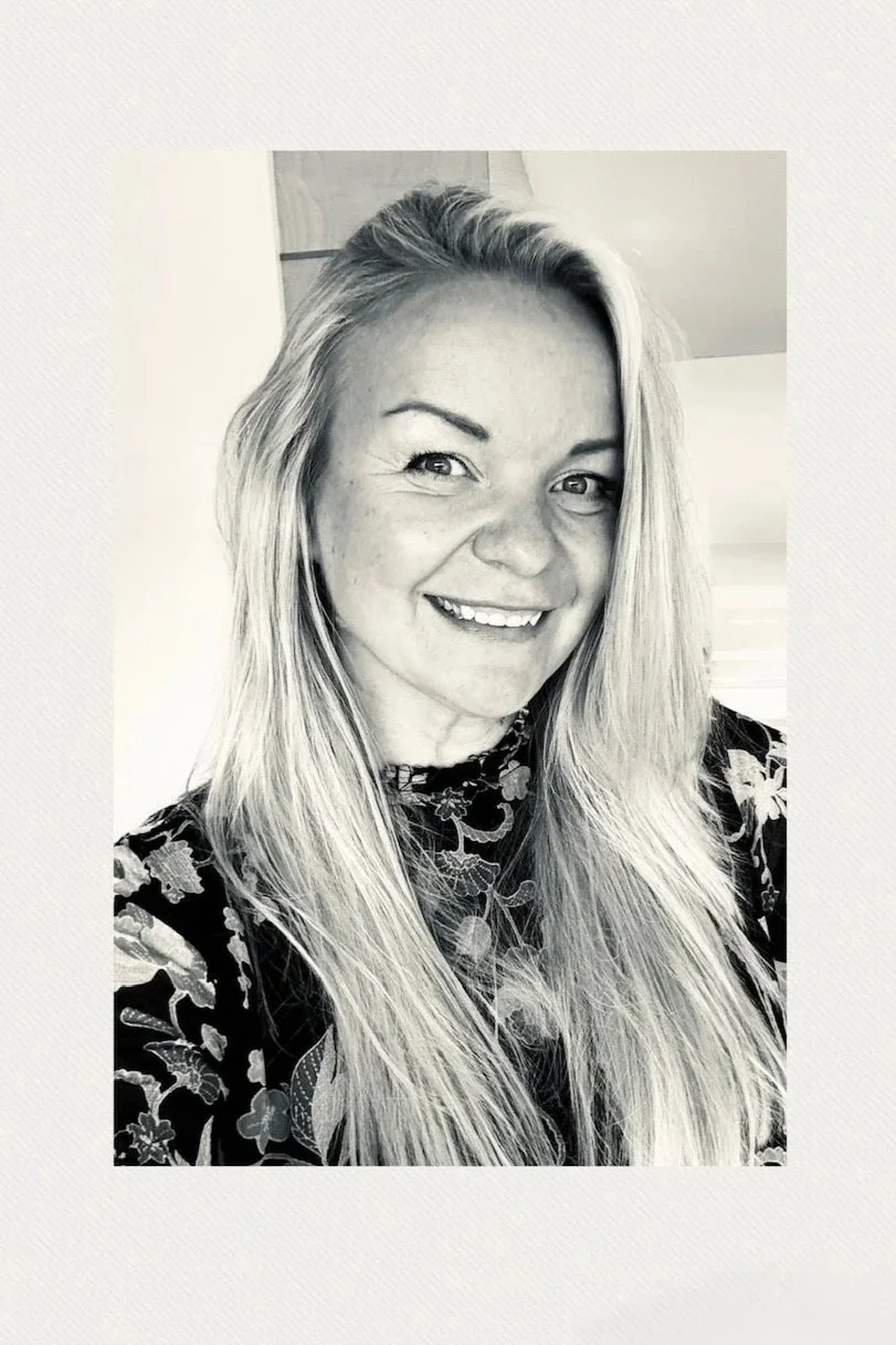 Black and white portrait of a smiling woman with long blonde hair wearing a floral-patterned top.