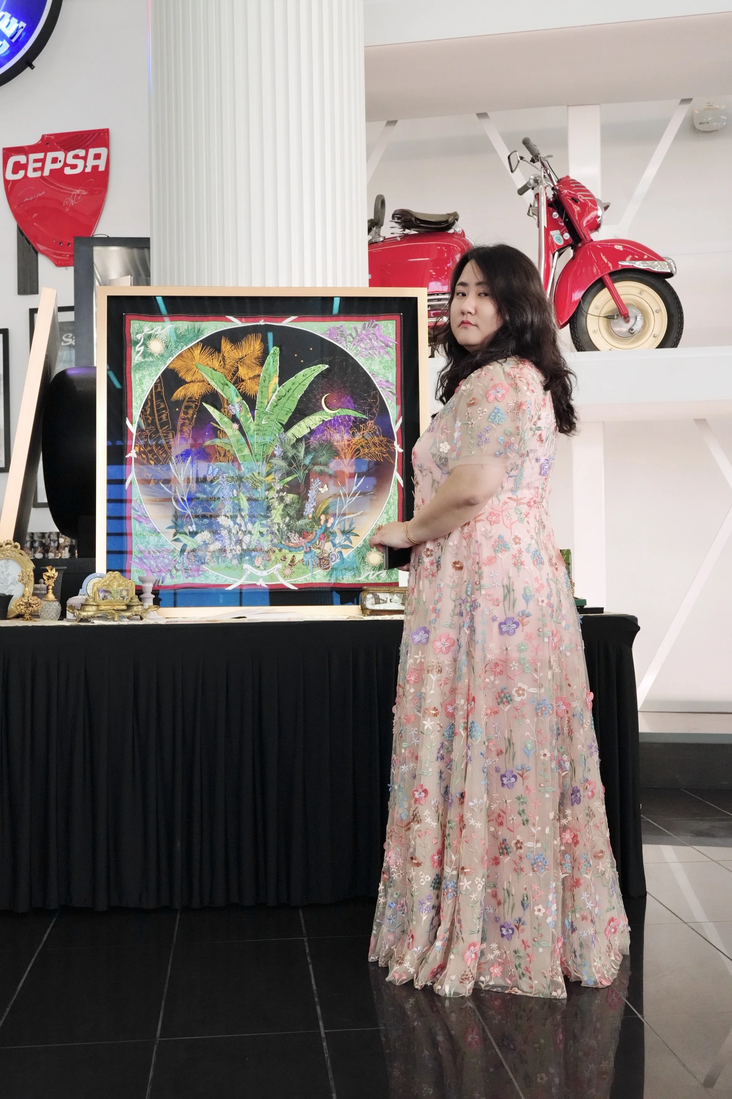 A woman with long dark hair wearing a long, sheer, floral dress standing next to a colorful framed artwork that depicts a tropical scene with palm trees and fireworks, in an art gallery with a red vintage motorcycle and a white wall in the background.