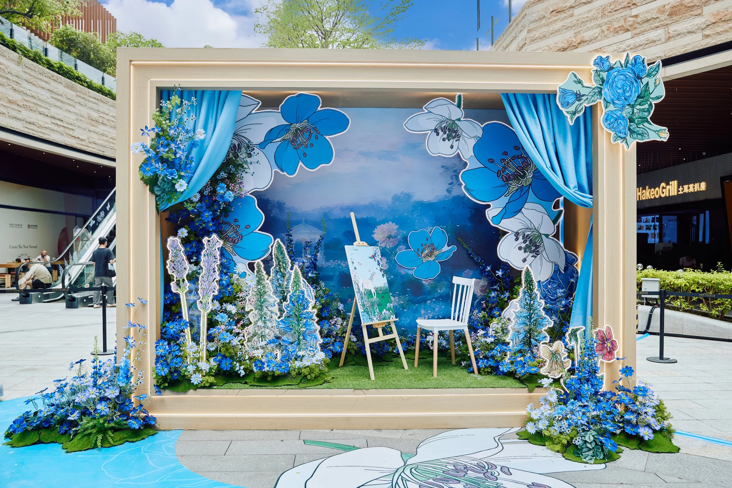 An outdoor decorative display featuring blue floral illustrations and cutouts, with blue curtains and a backdrop of a scenic sky and mountain landscape.