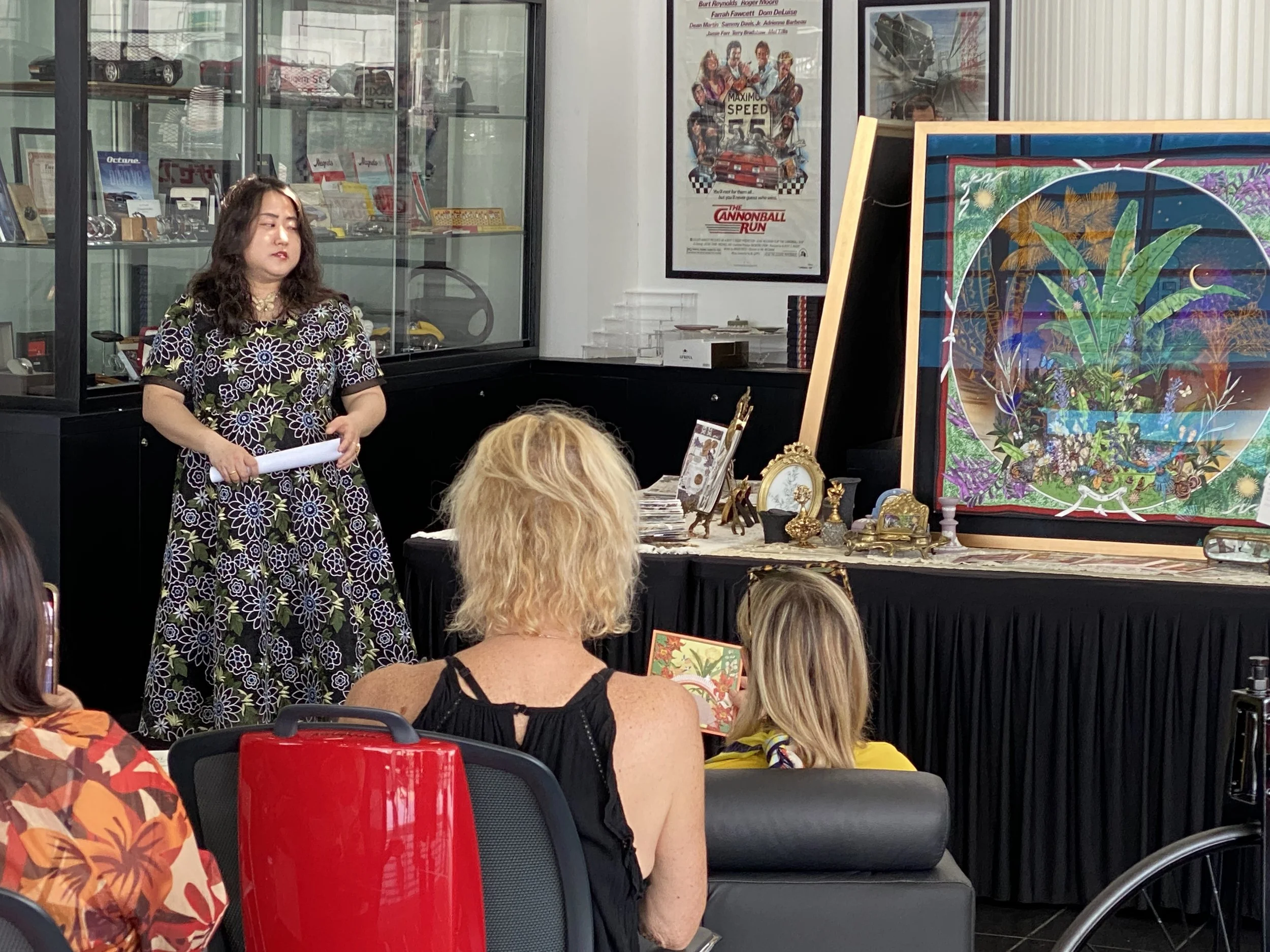 A woman in a floral dress standing in front of a small audience at an art auction or presentation. Behind her, there is a glass display case with model cars, posters on the wall including a 'The Cannonball Run' poster, and a colorful artwork on an easel. Several audience members are seated, watching her.