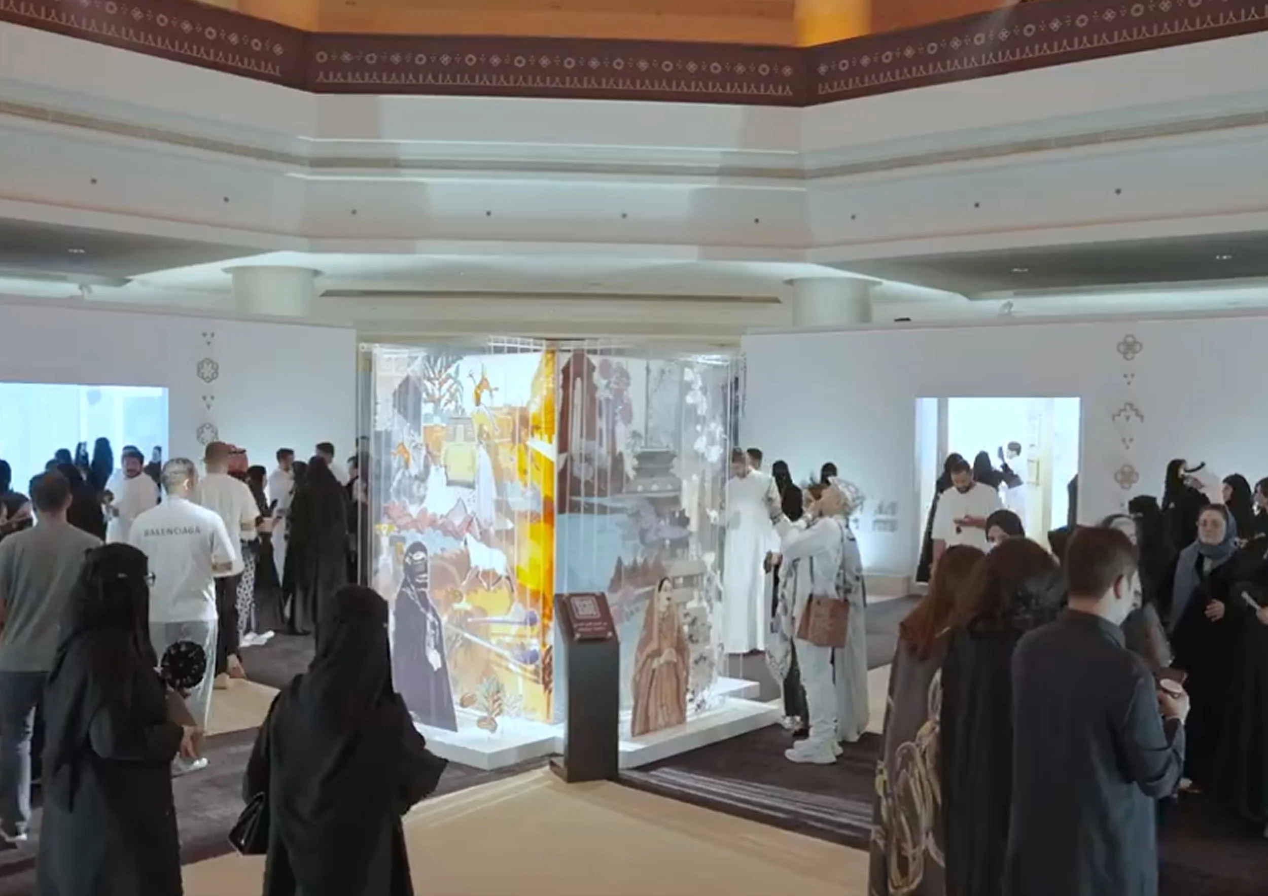 People gathered inside a building, possibly a gallery or museum, with colorful artwork on display and decorative patterns on the walls.