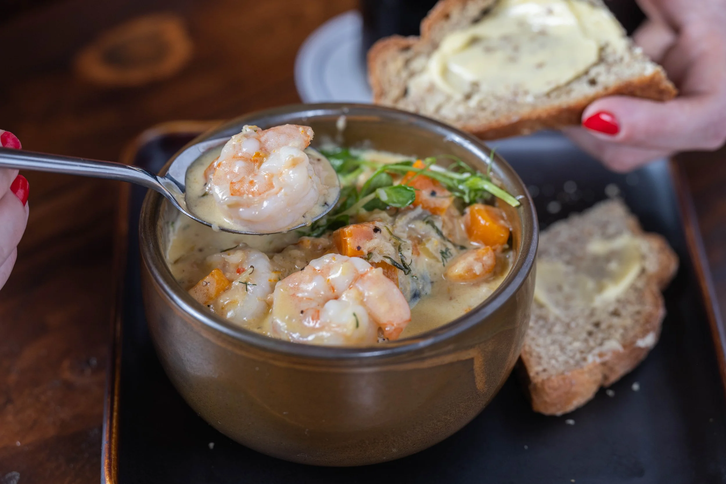 A bowl of creamy shrimp and vegetable stew with a spoon lifting a piece, accompanied by slices of bread with butter on a dark tray.